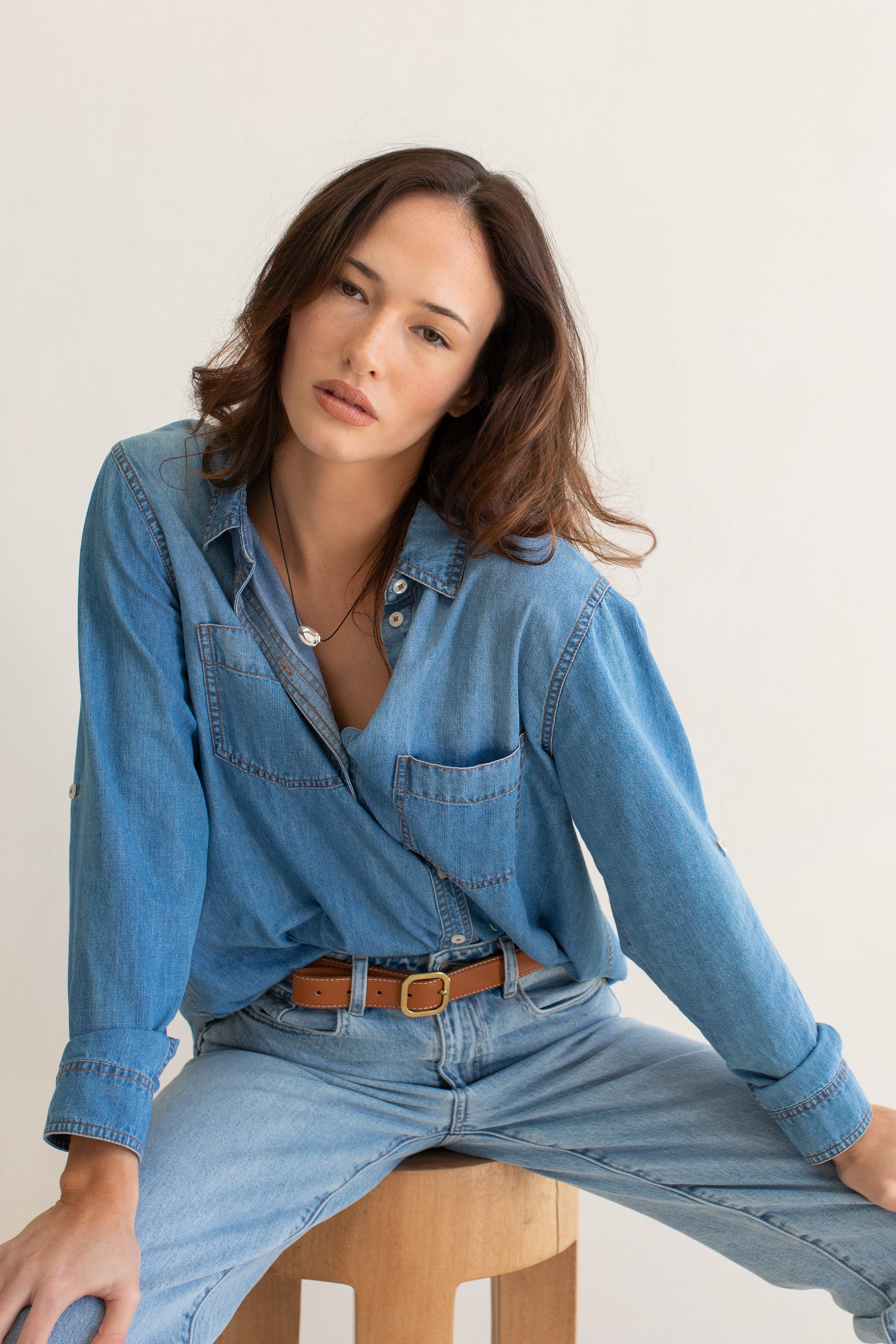Woman wearing a blue Hut denim oversized boyfriend shirt and jeans sitting on a wooden stool against a white background