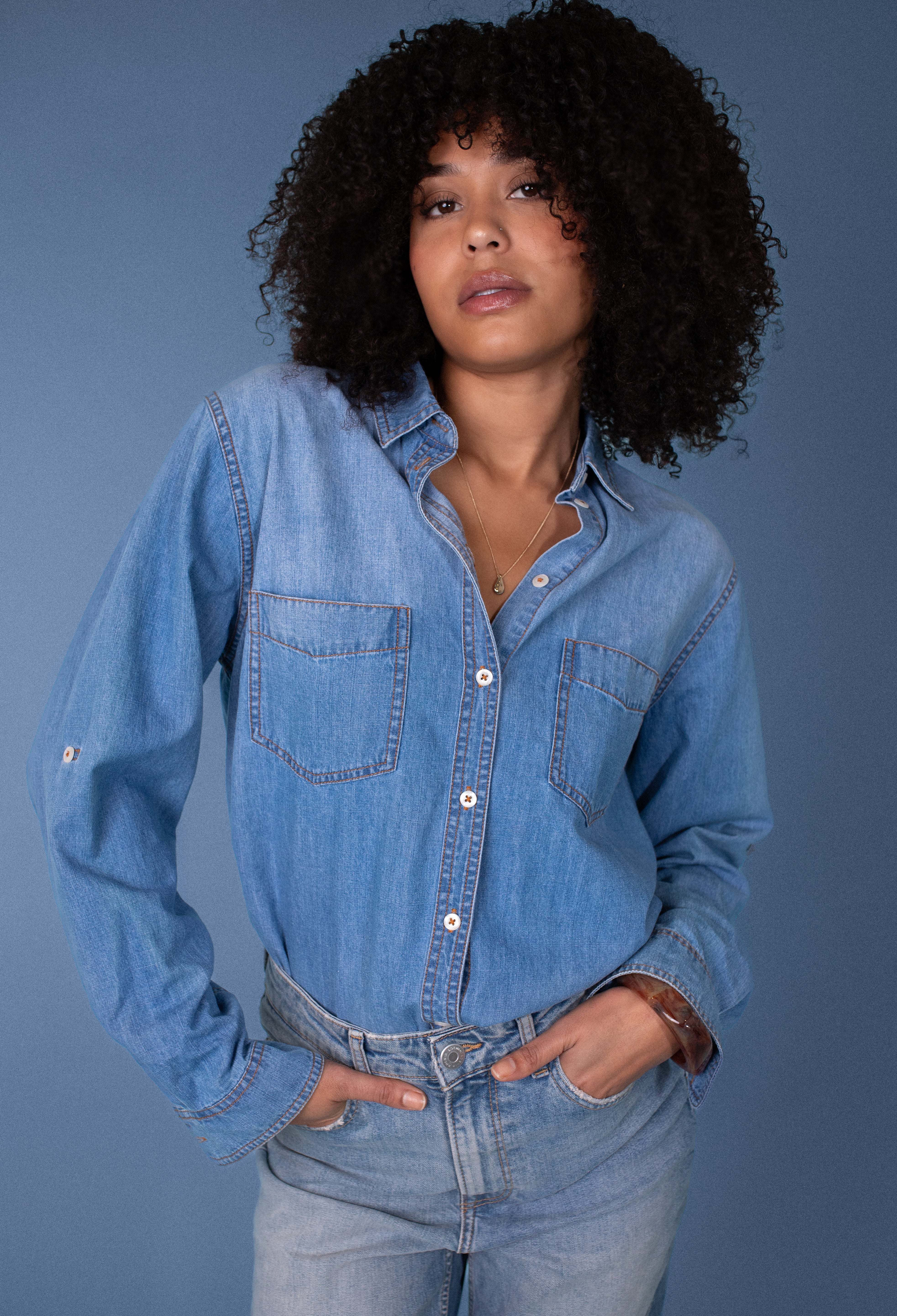 Woman wearing a blue Hut denim shirt and jeans against a plain background