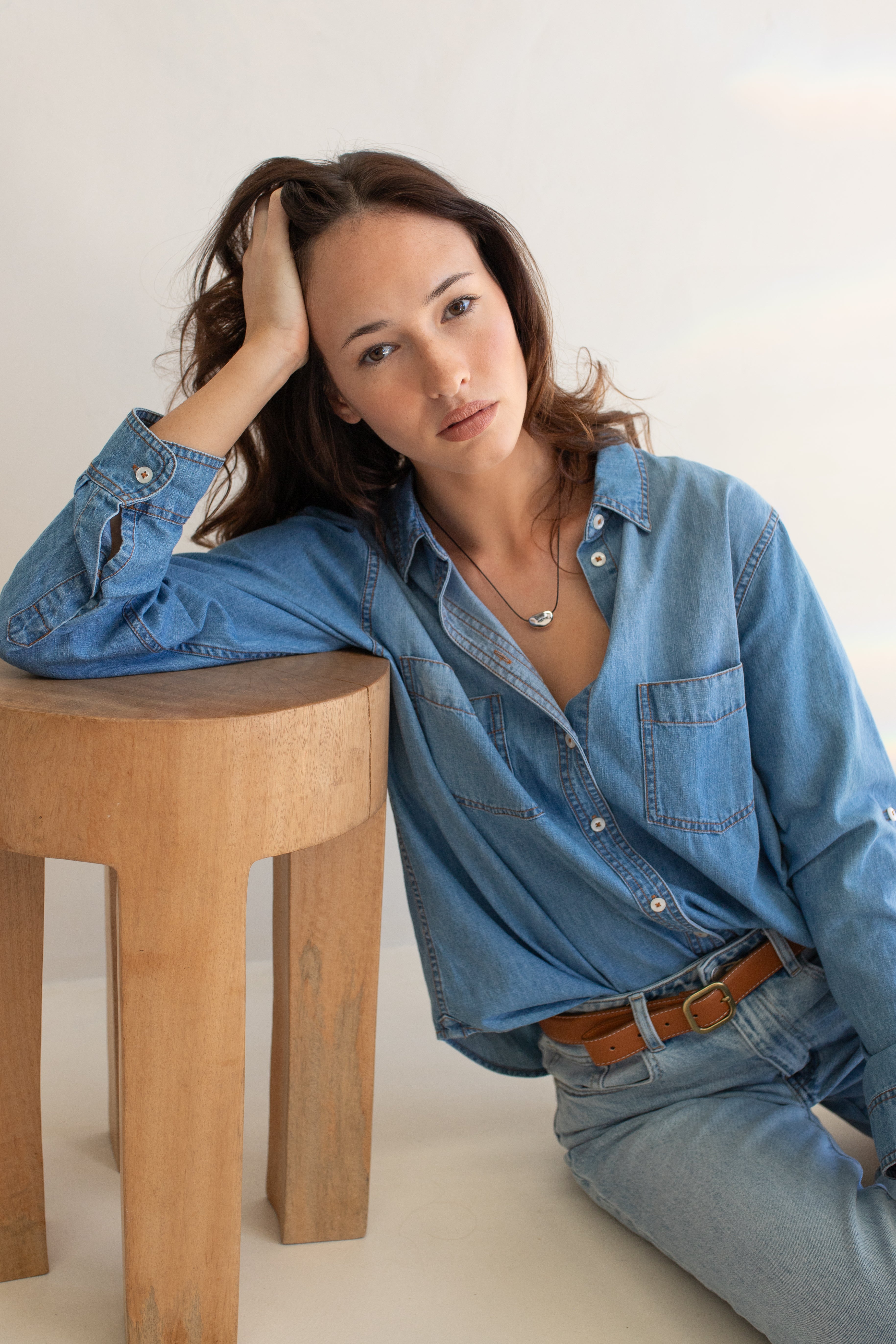 Woman wearing a blue denim shirt and jeans sitting on a wooden stool against a white background
