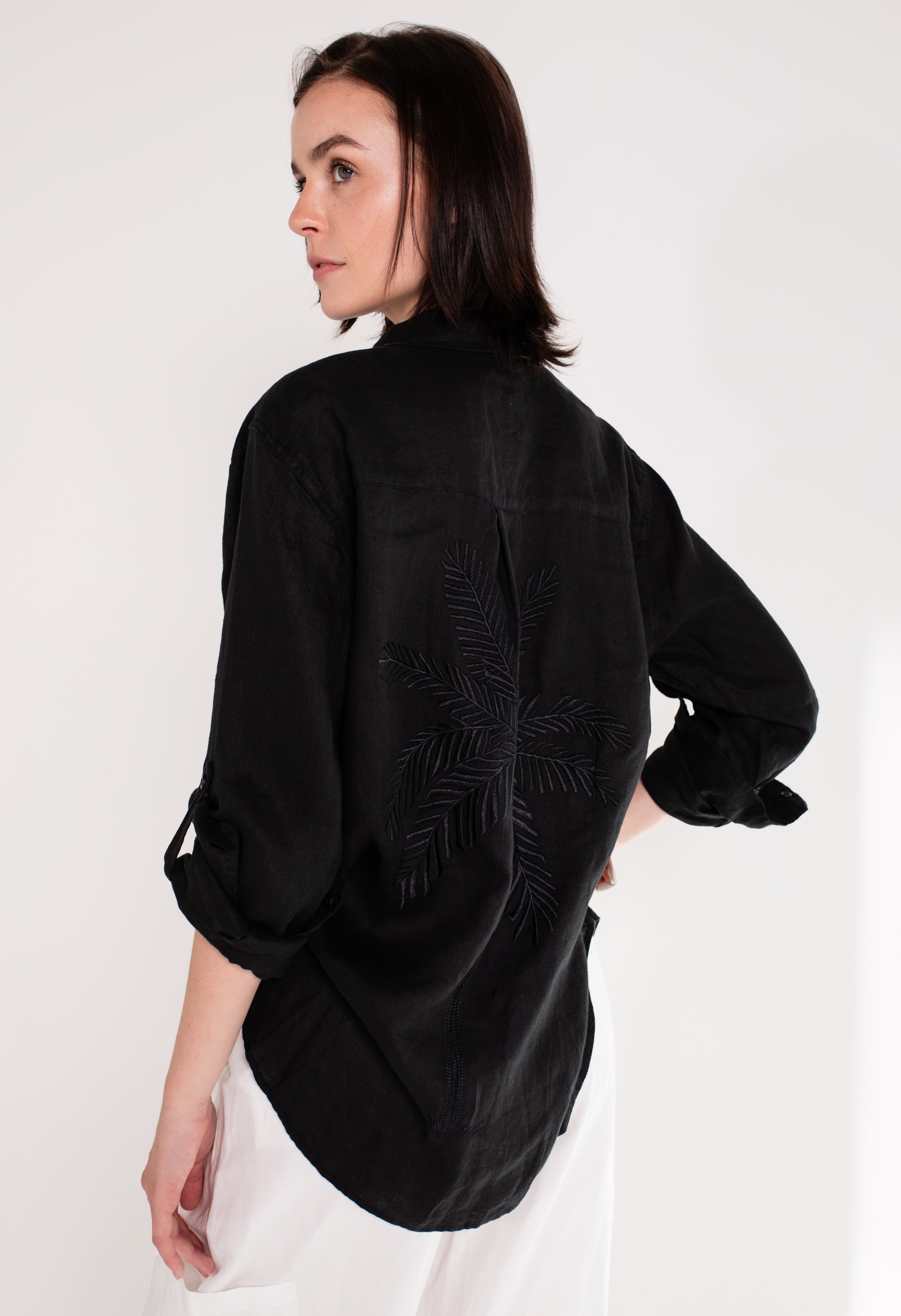 A model wearing Hut clothing black boyfriend linen shirt with an embroidered palm tree on the back, standing against a white background.