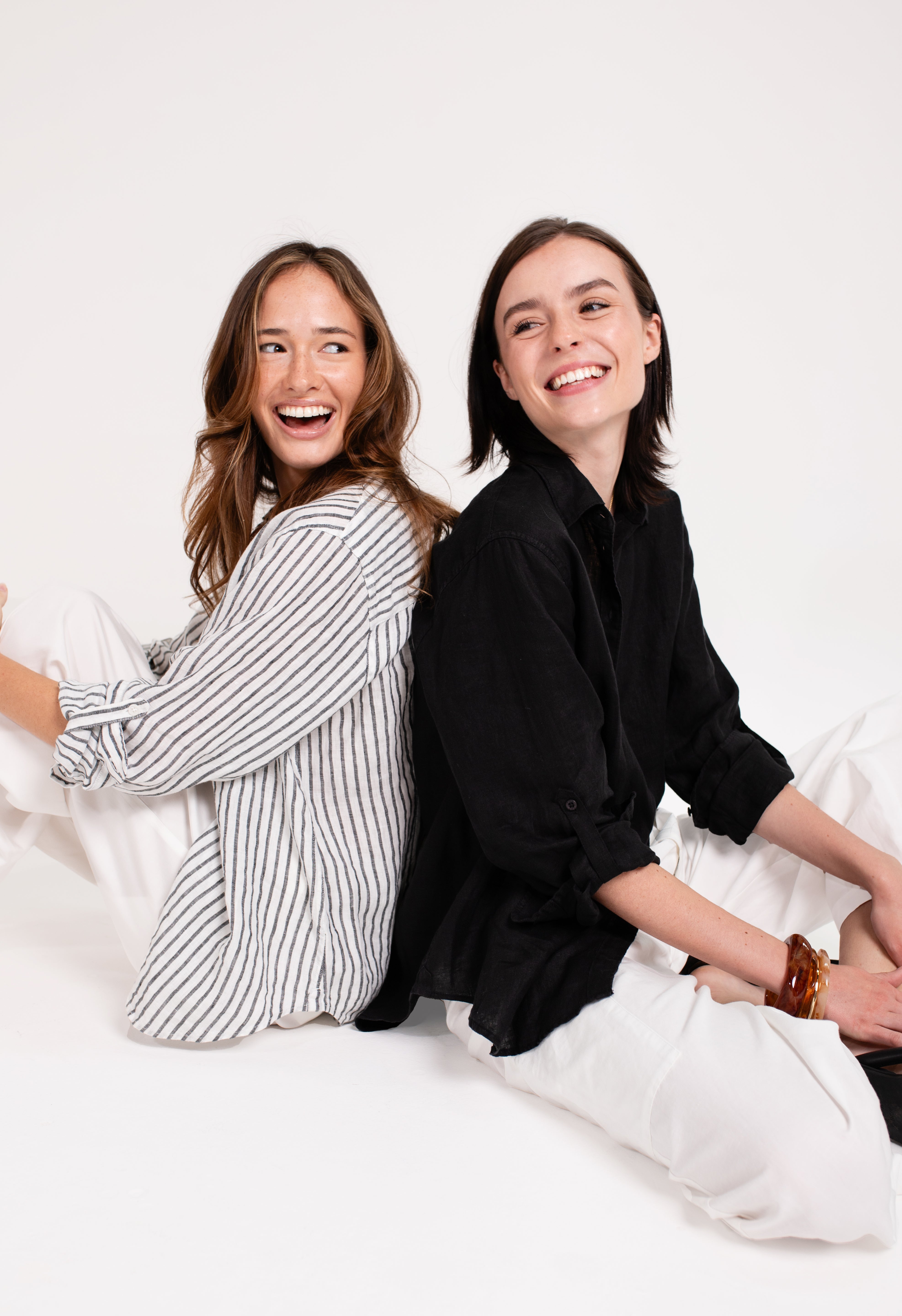 Two models back to back laughing wearing Hut Black Embroidered Palm Boyfriend Linen Shirt and Hut Black and White Stripe Boyfriend Linen Shirt