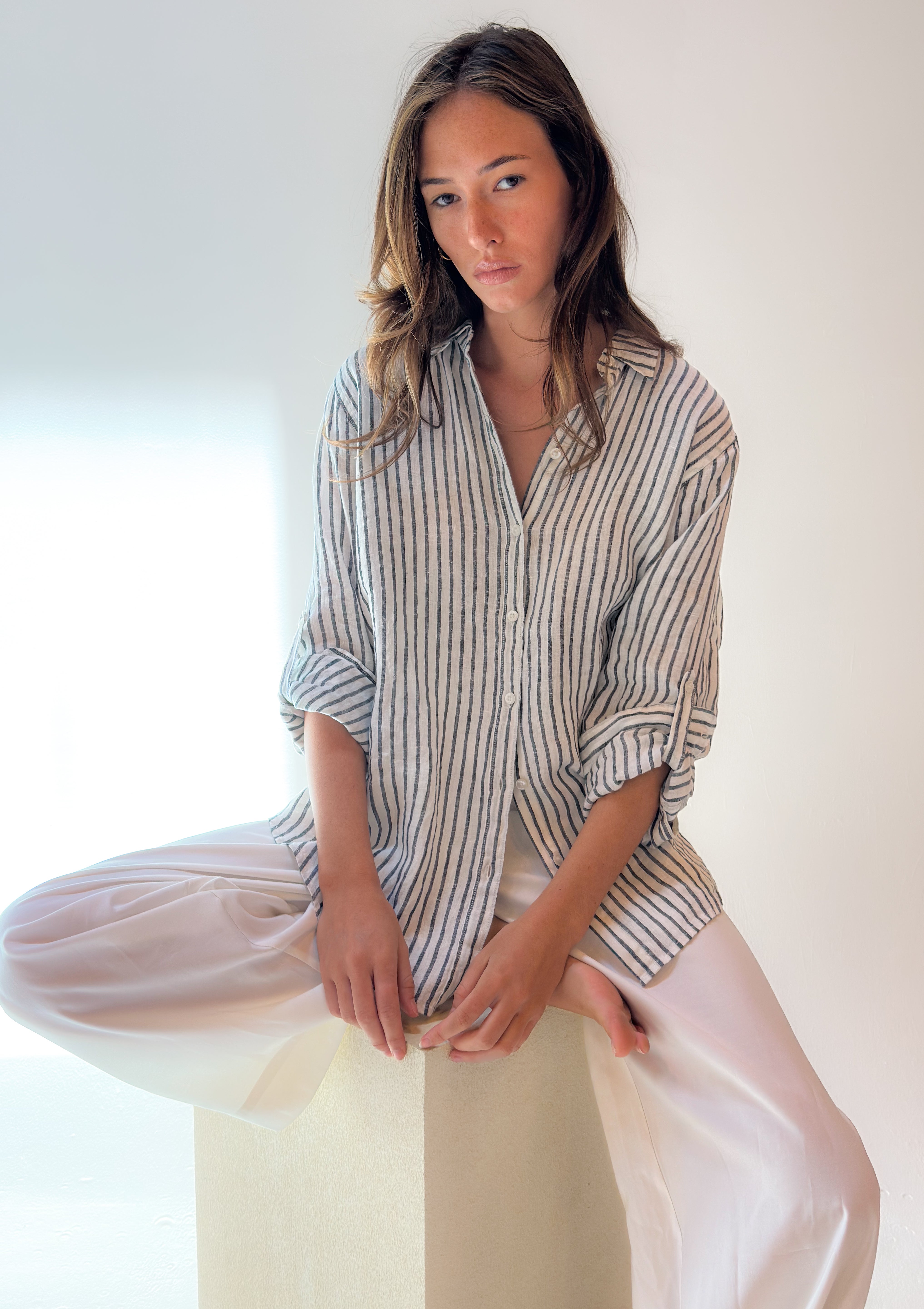 A woman sitting on a pedestal wearing a striped Hut clothing boyfriend linen shirt with button-up sleeves and a relaxed fit. Made from organic linen