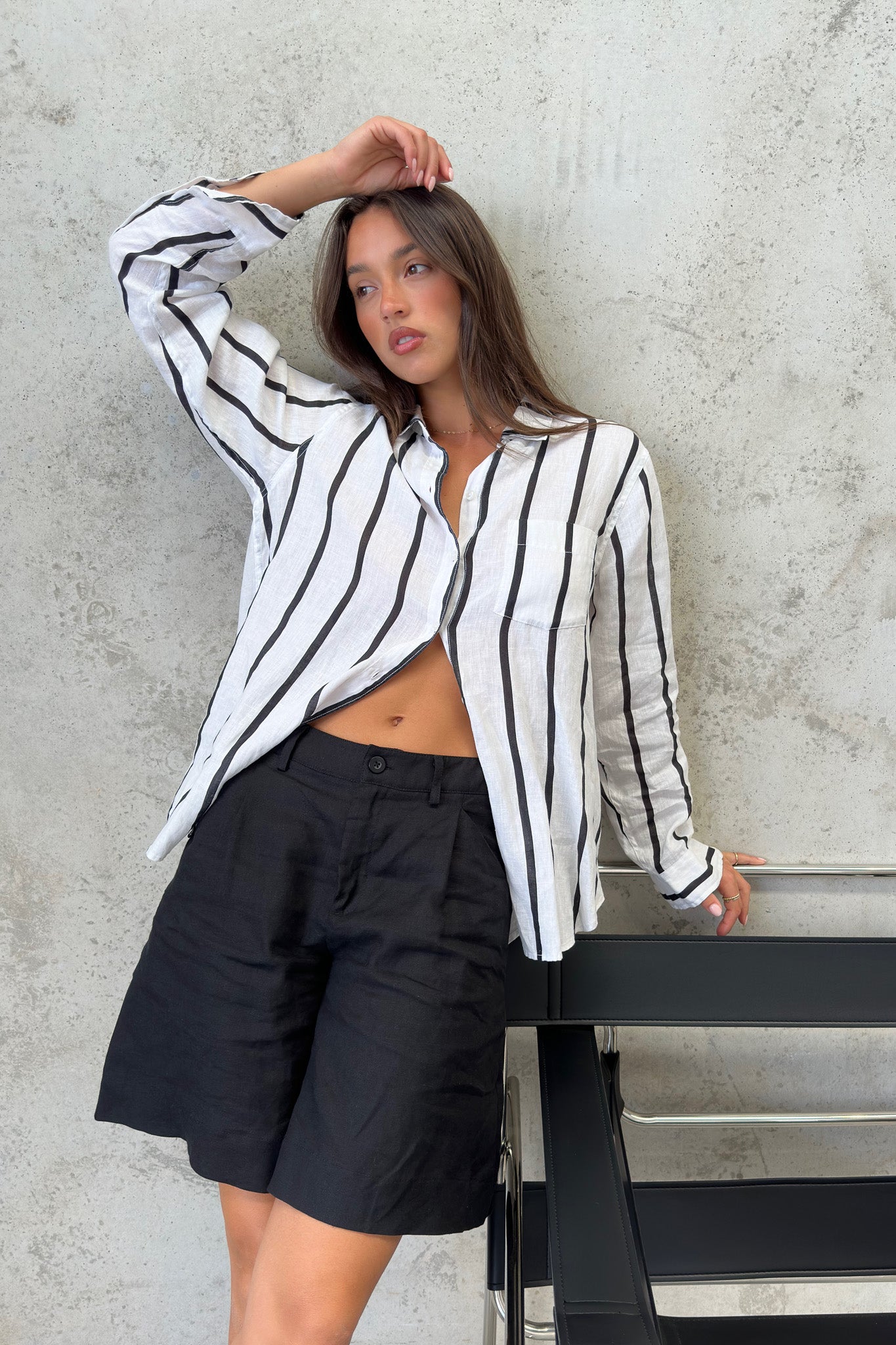 A woman wearing a black and white striped Hut boyfriend linen shirt with French seams and a scoop hem, paired with black shorts.
