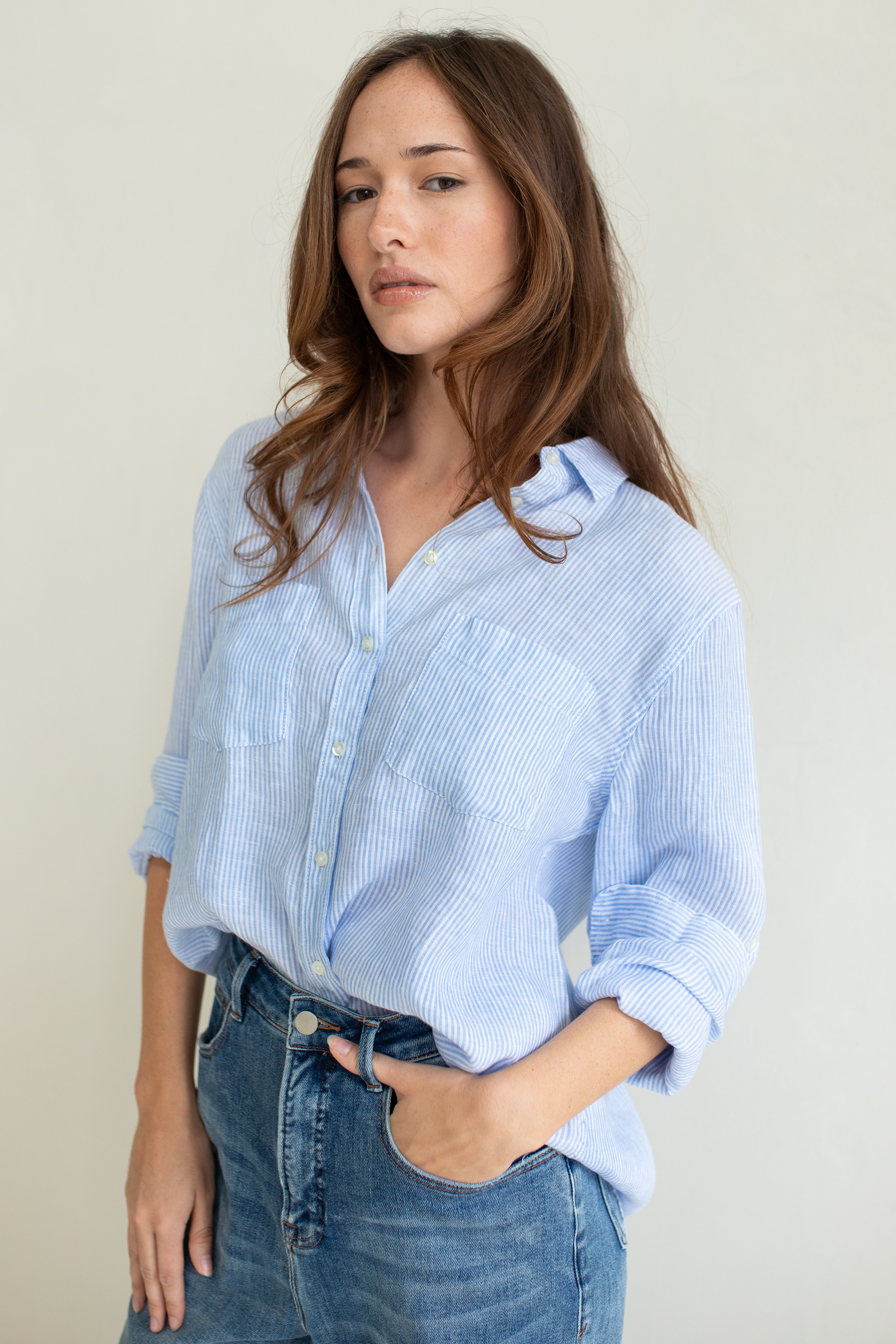 A woman wearing a baby blue striped linen boyfriend shirt with long sleeves and a scoop hem, paired with blue jeans.