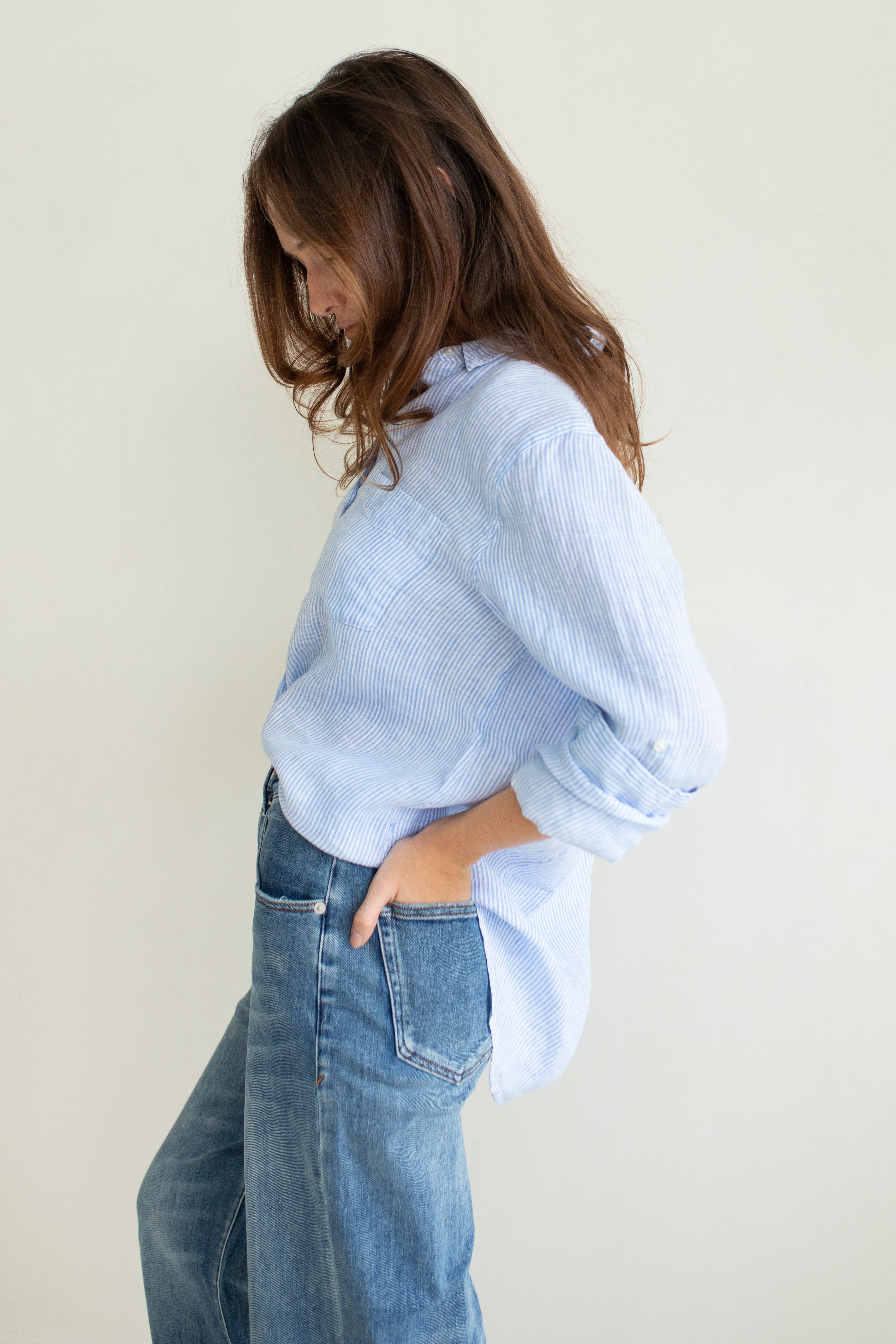 Side profile of Hut Clothing Baby Blue Stripe Boyfriend Linen Shirt with models in hand in back of jean pocket and tab sleeves rolled up