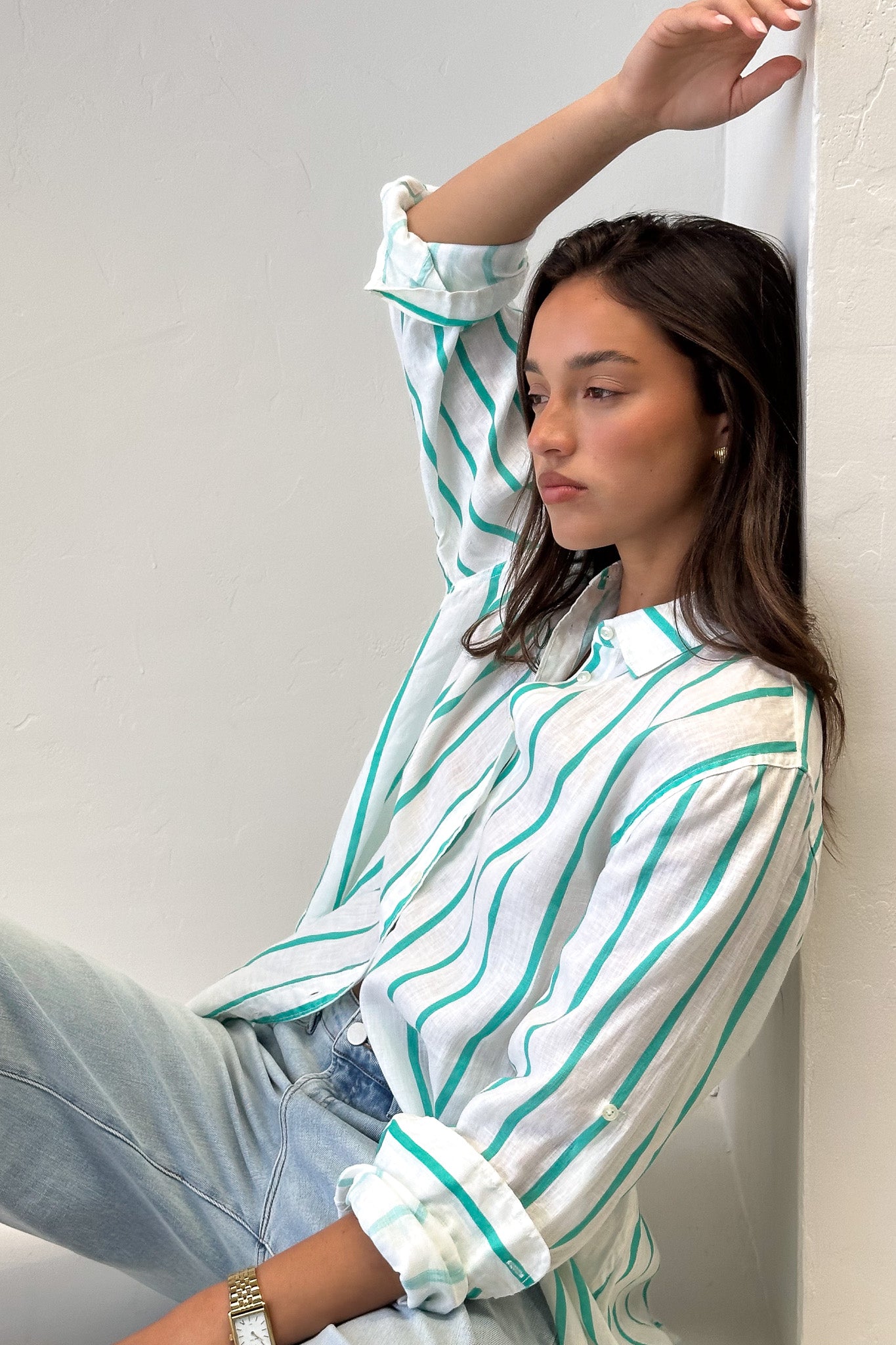 A woman sitting against a wall wearing a striped boyfriend linen shirt in green and white with rolled-up sleeves, light wash denim jeans