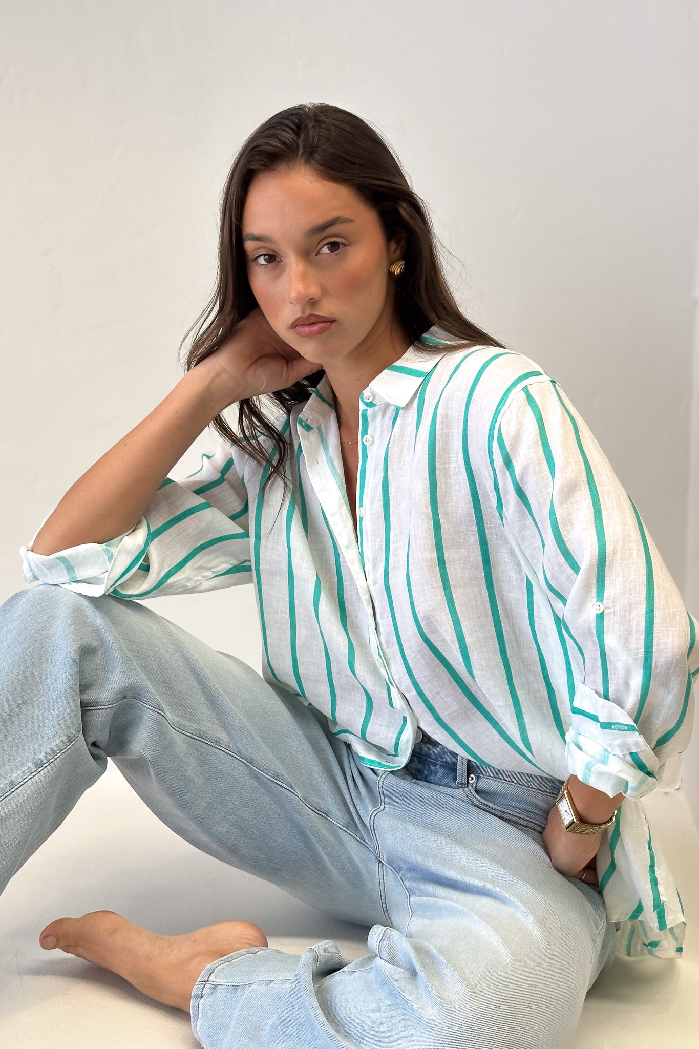 Model sitting down wearing Bermuda Green Stripe Boyfriend Linen shirt with light wash denim jeans