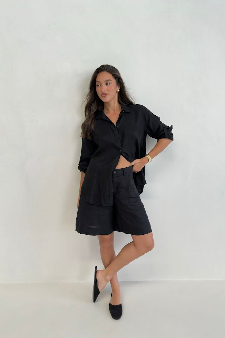 A model standing against a white background, wearing a black boyfriend linen shirt with button-up sleeves and black shorts.