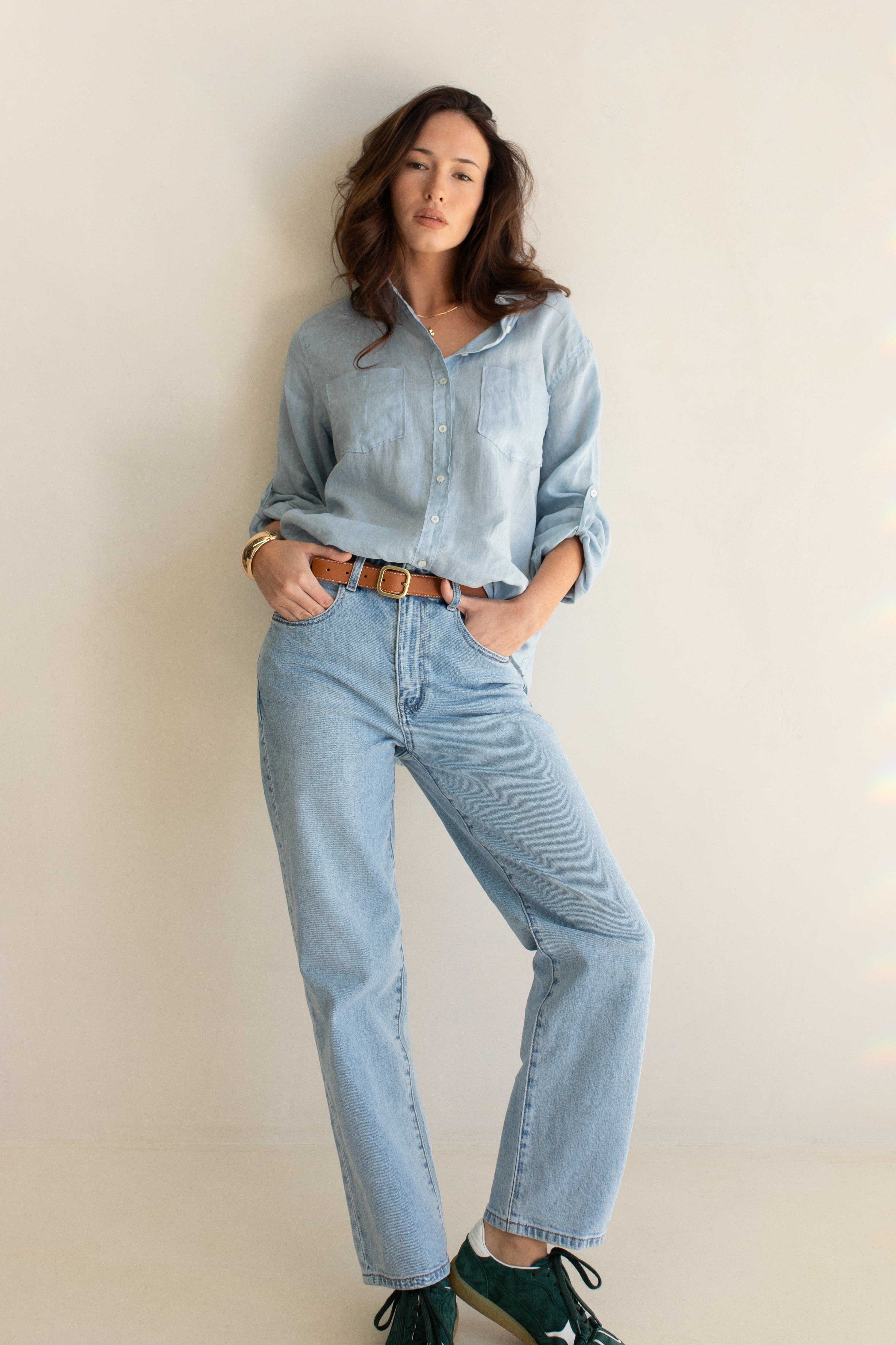 Woman wearing a light blue linen boyfriend shirt and jeans against a beige wall