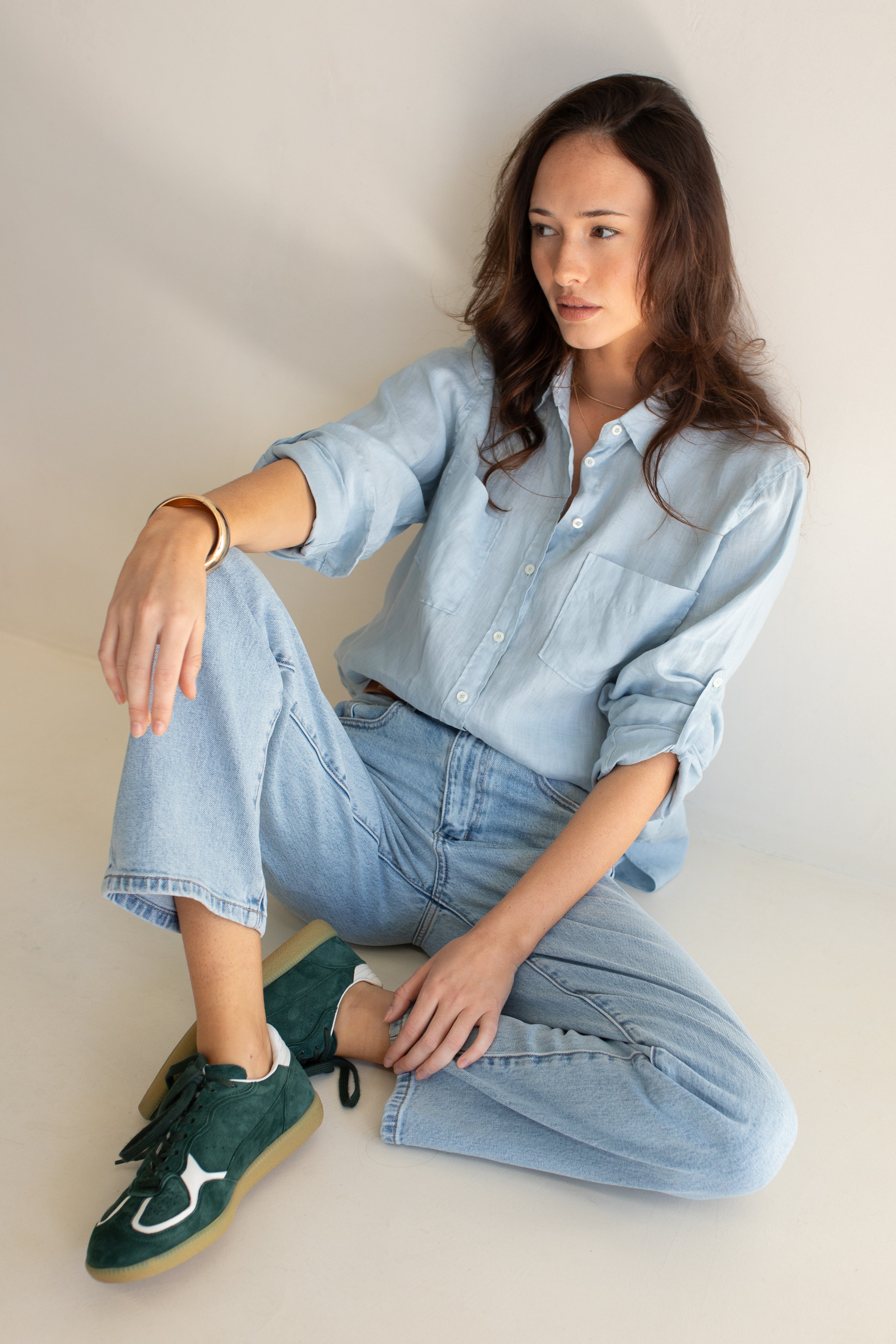 Model sitting down wearing Hut Clothing Breeze Blue Boyfriend Linen Shirt with light denim jeans and green sneakers
