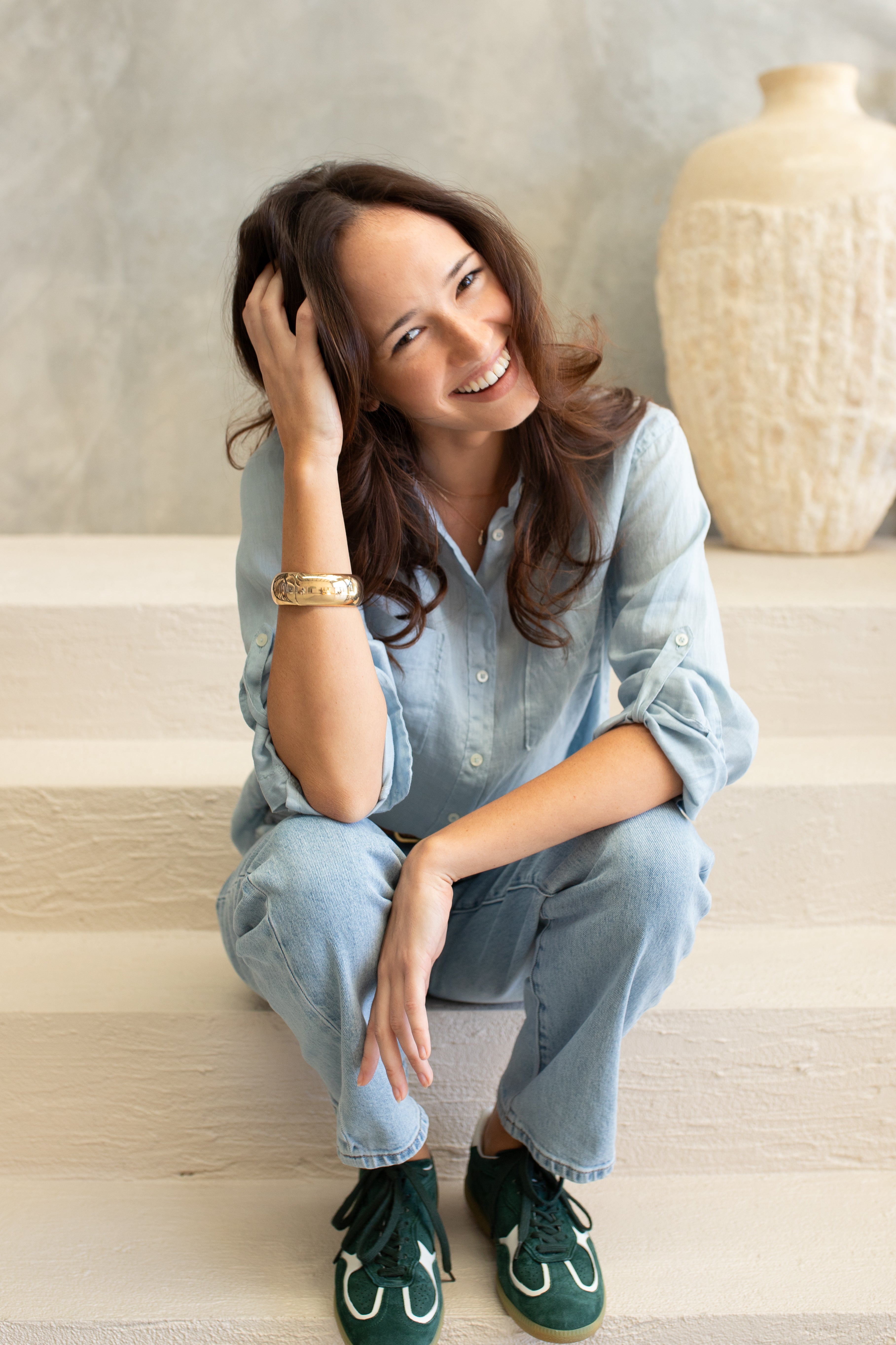 Woman sitting on a staircase wearing a light blue linen boyfriend shirt and jeans, smiling.