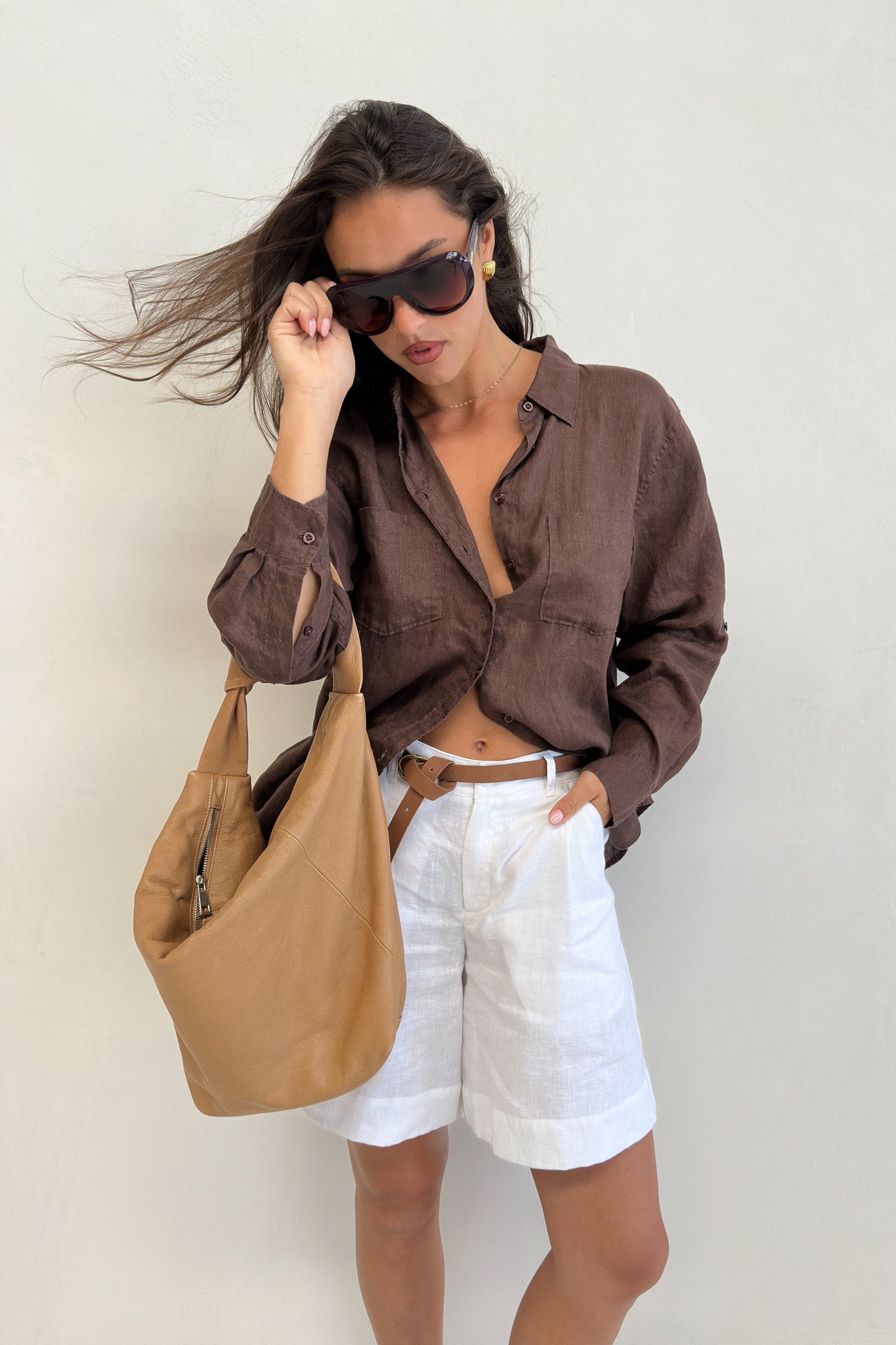 Model wearing Hut Clothing Chocolate Martini Boyfriend Linen Shirt with wind in her hair and a tan leather bag with white linen shorts