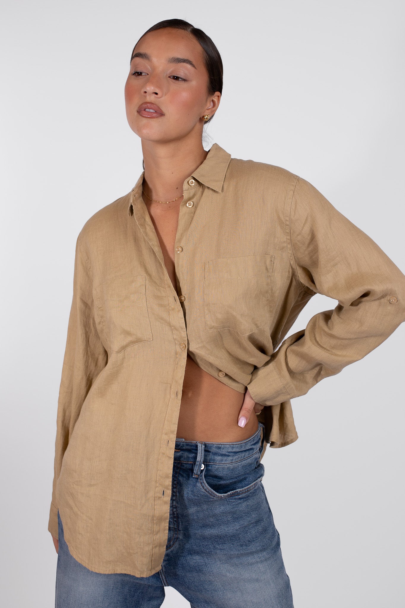 A model wearing an oversized beige linen shirt and blue jeans. The shirt is buttoned at the top but unbuttoned at the bottom, tab sleeves, two chest pockets in the Hut Boyfriend Fit style