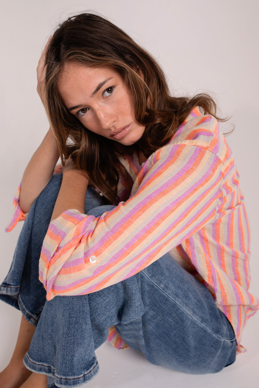 A woman sitting wearing a striped Hut boyfriend linen shirt in multi colors with button-up sleeves and a collar.