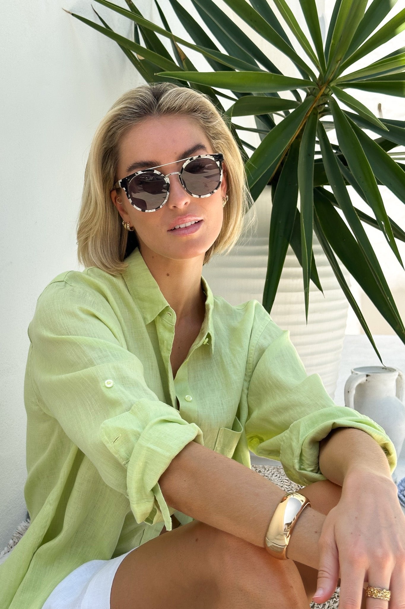 A woman wearing a green linen boyfriend shirt with rolled-up sleeves, paired with white shorts, sitting beside a potted plant.