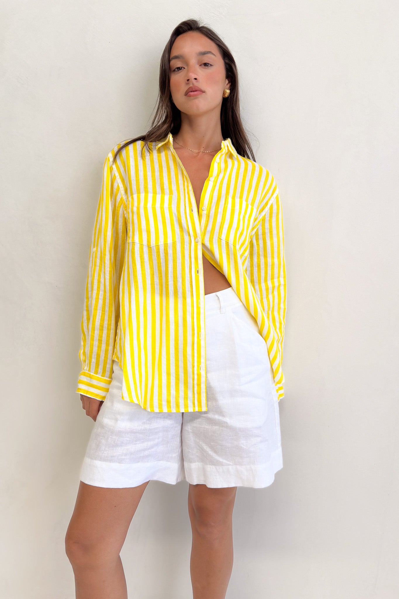 A woman wearing a Hut Clothing yellow and white striped linen boyfriend shirt with long sleeves and two chest pockets, paired with white shorts.