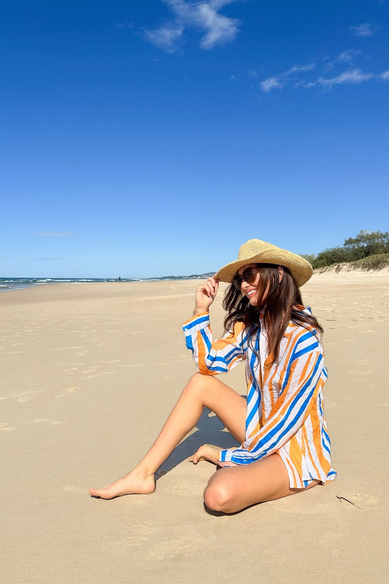 Woman sitting on a sandy beach wearing an orange and blue striped linen oversized shirt and hat.