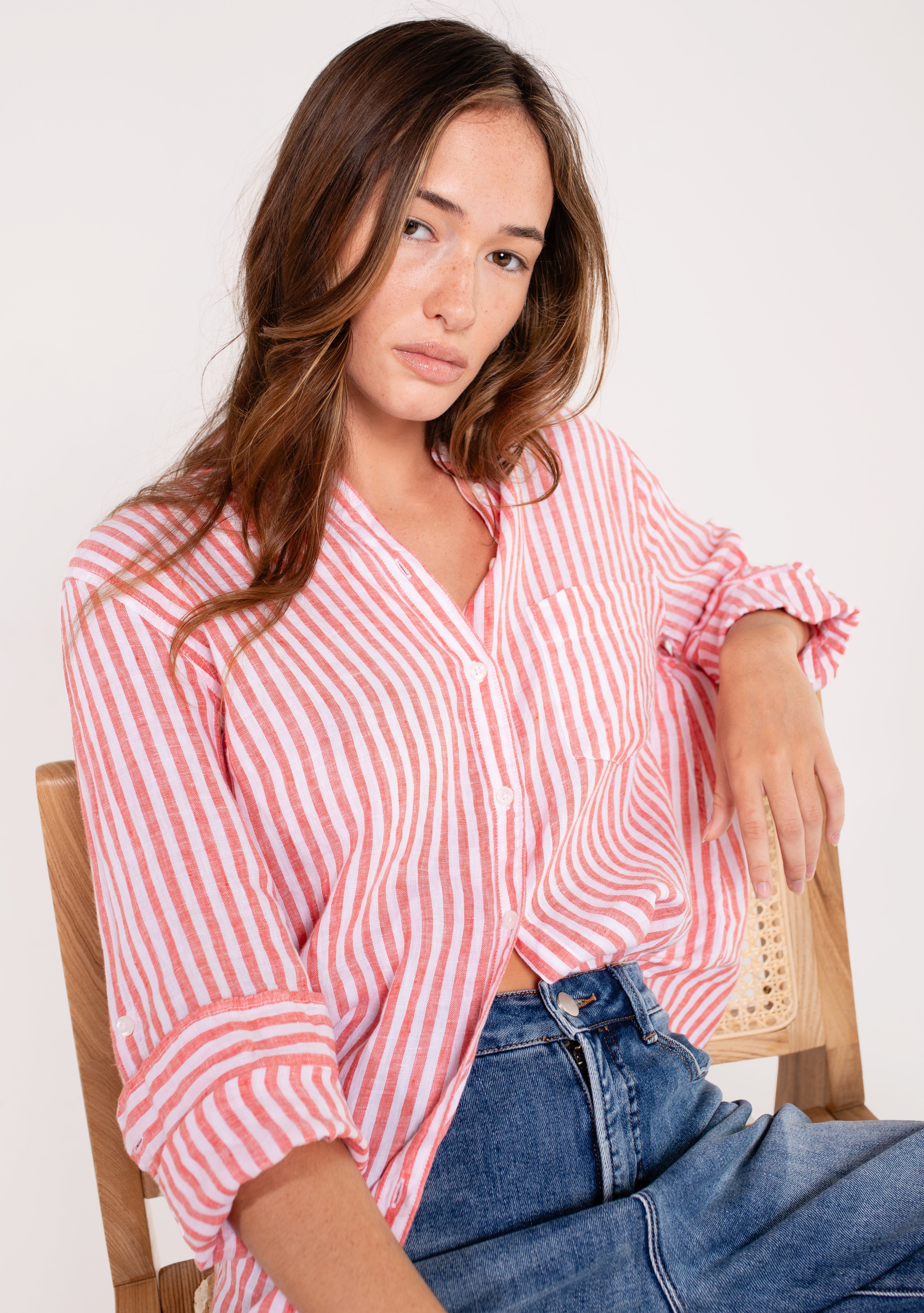 A woman sitting and wearing a Hut red and white striped boyfriend linen shirt with button-up sleeves and a scoop hem, paired with blue jeans.
