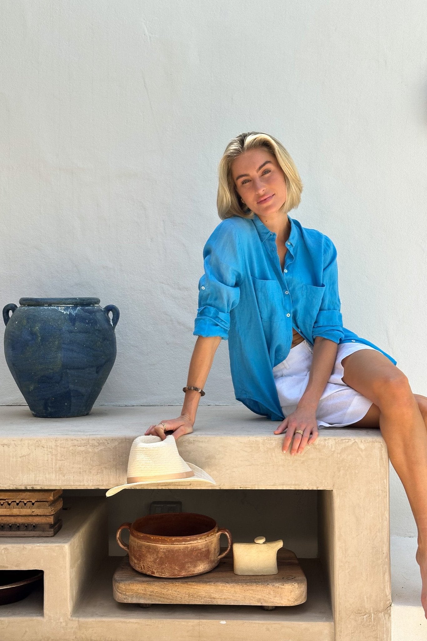 A woman sitting on a stone bench wearing a blue linen shirt with white shorts, accessorized with a watch and earrings.