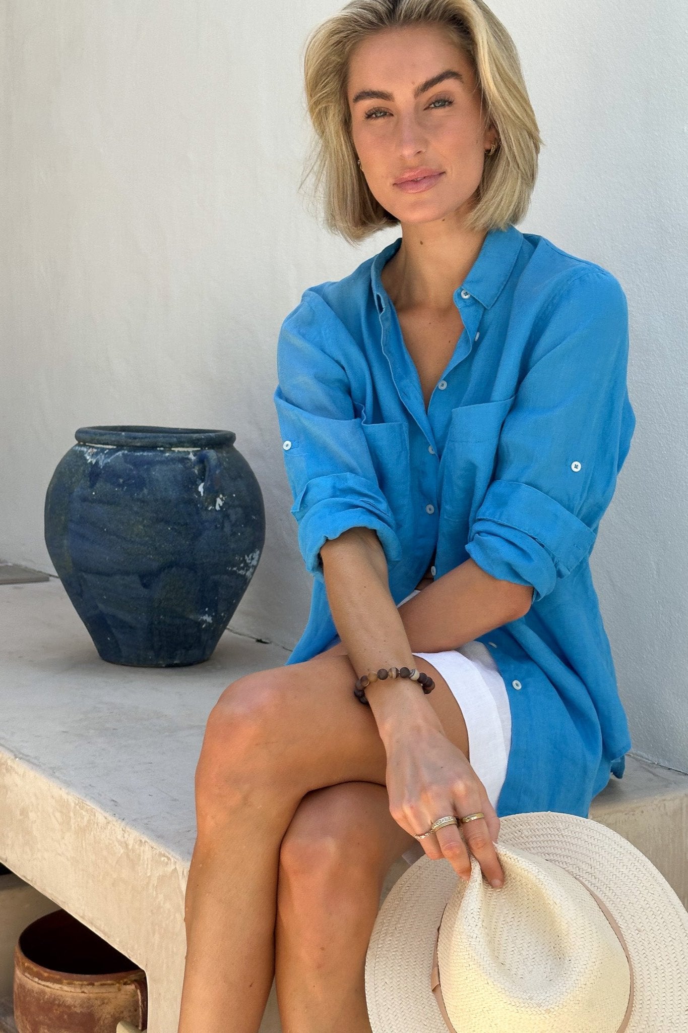 Model sitting on stone bench wearing Hut Reef Blue Boyfriend Linen Shirt holding a hat