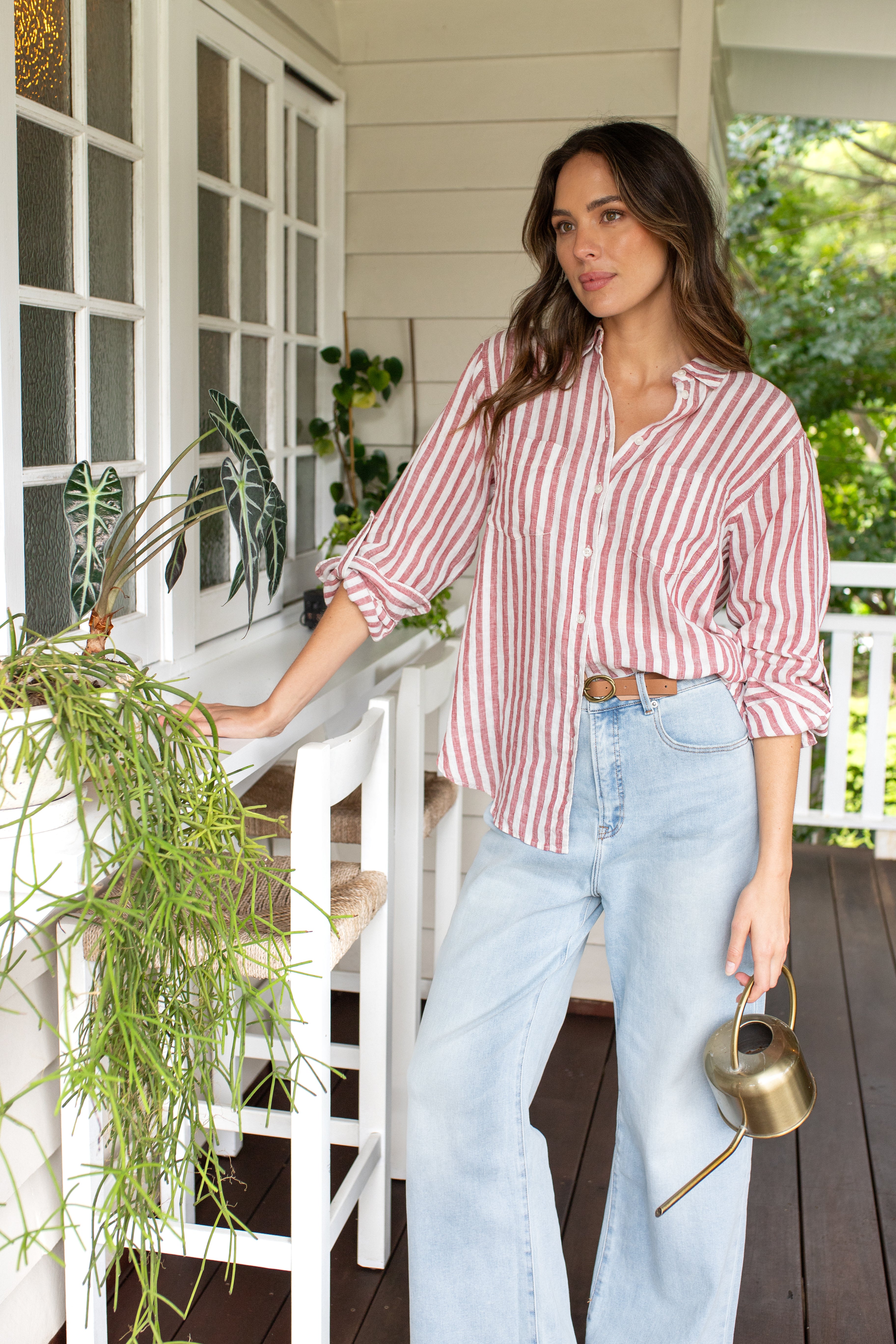 A woman standing on a porch wearing a Hut Rhubarb striped boyfriend linen shirt with long sleeves and light blue jeans. She is holding a watering can and there are plants in the background.