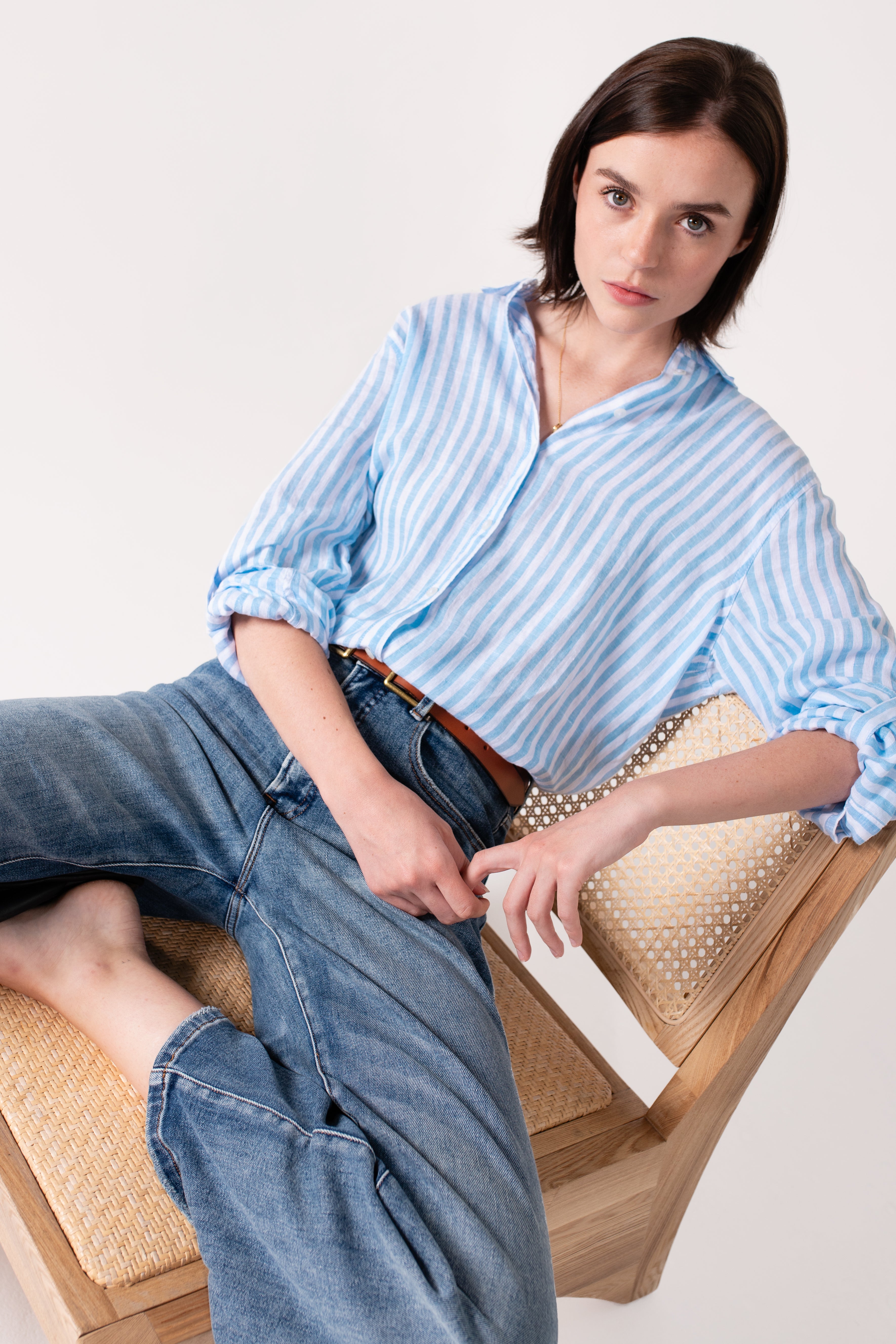 Model wearing a Hut Clothing sky blue yarn dyed stripe boyfriend linen shirt with rolled-up sleeves, paired with blue jeans, sitting on a chair.