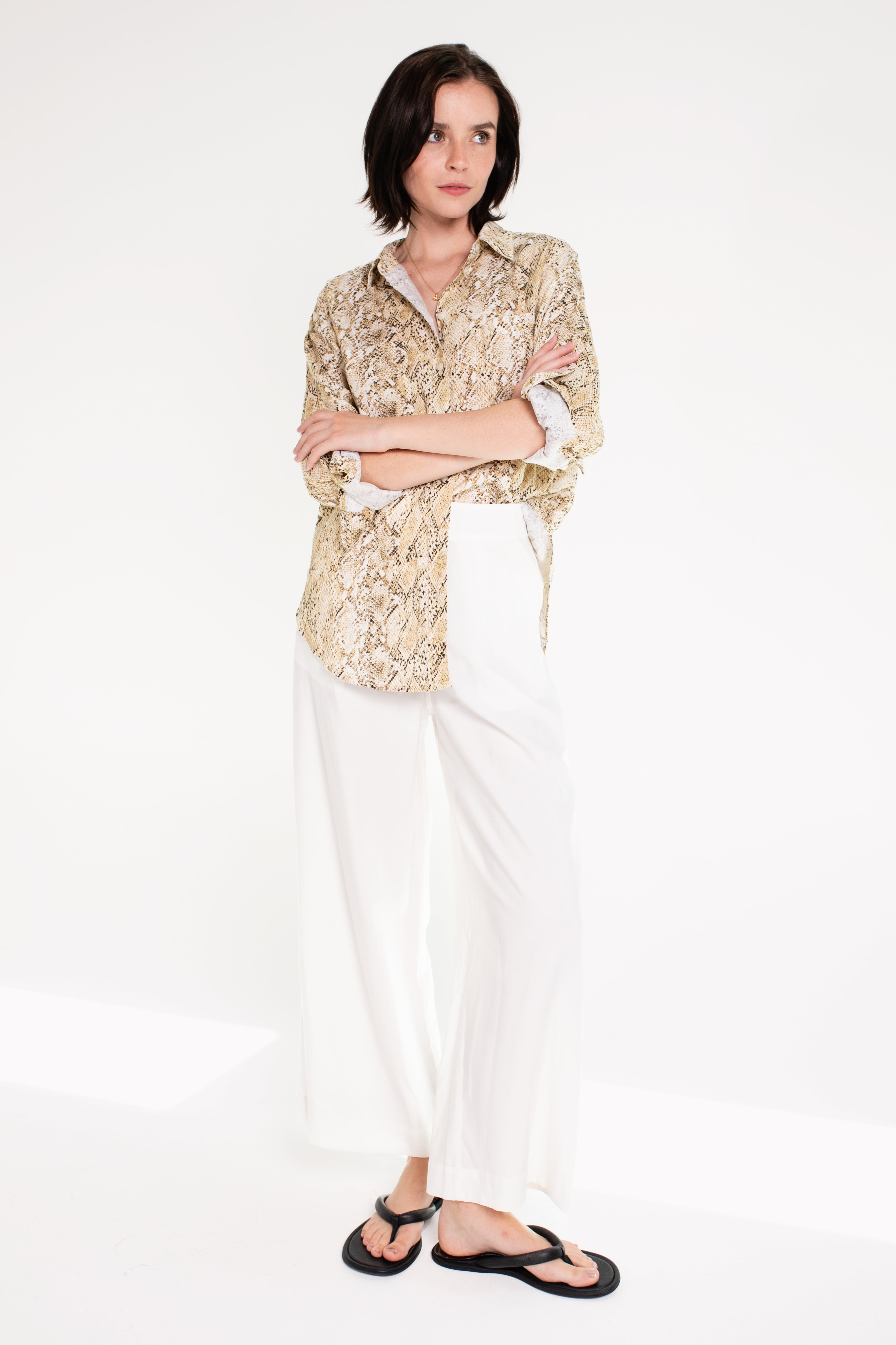 A woman wearing a Hut snake skin print boyfriend linen shirt in an oversized fit, paired with white trousers, standing against a white background.