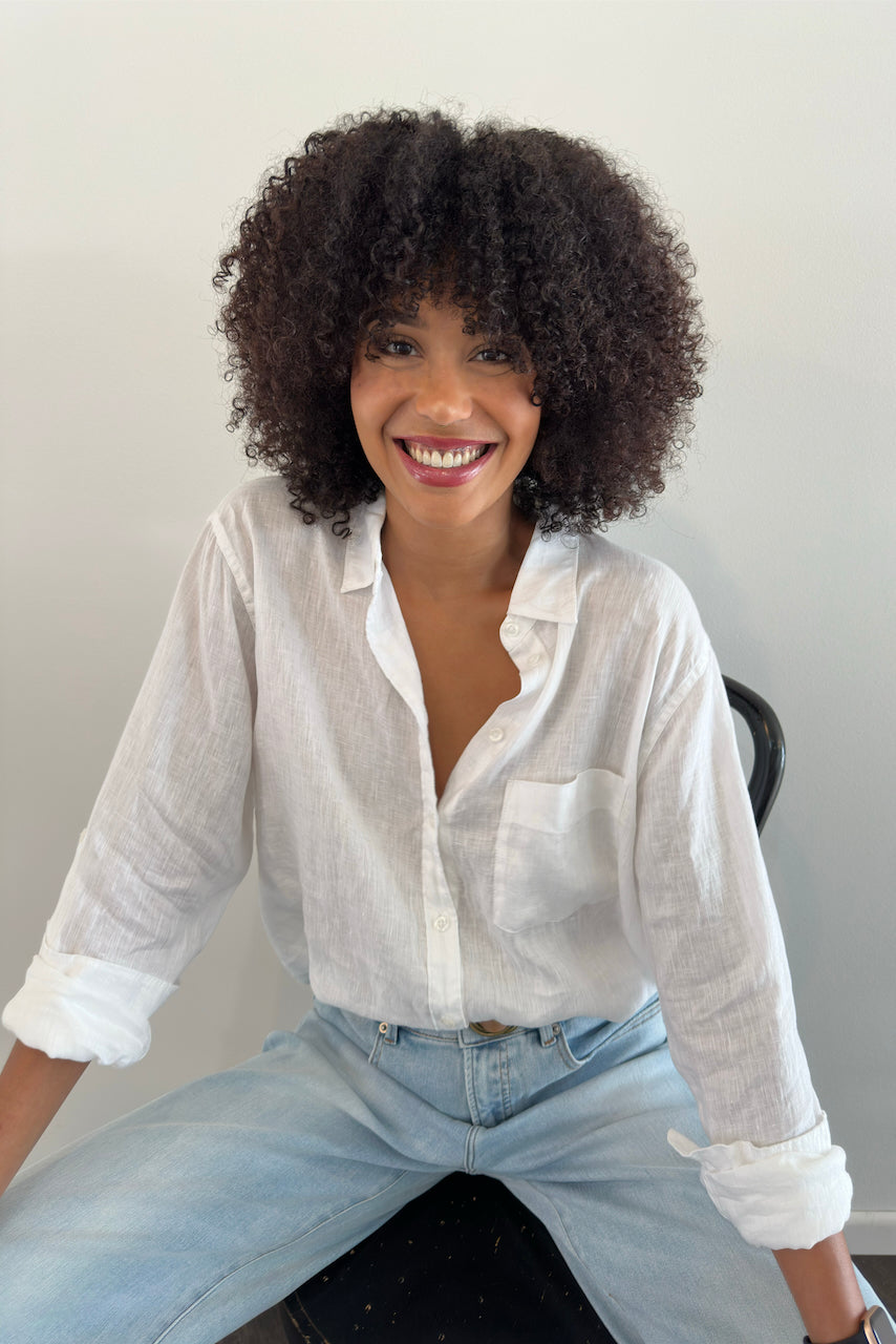A model smiling, wearing a white linen Hut clothing shirt with one chest pocket and light blue jeans.