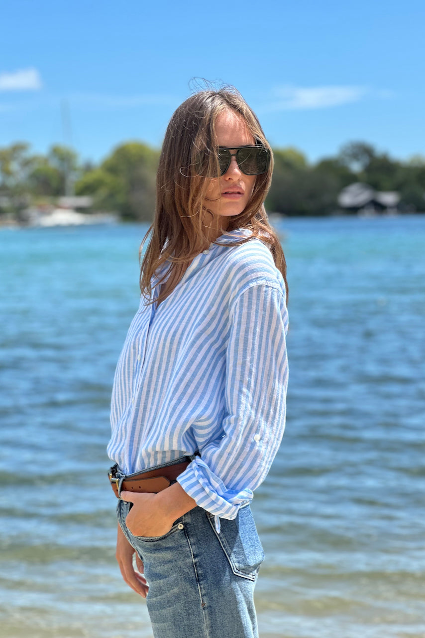 A woman standing by the water wearing a striped Hut Clothing boyfriend linen shirt in blue and white.