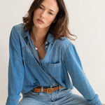 Woman wearing a blue Hut denim oversized boyfriend shirt and jeans sitting on a wooden stool against a white background