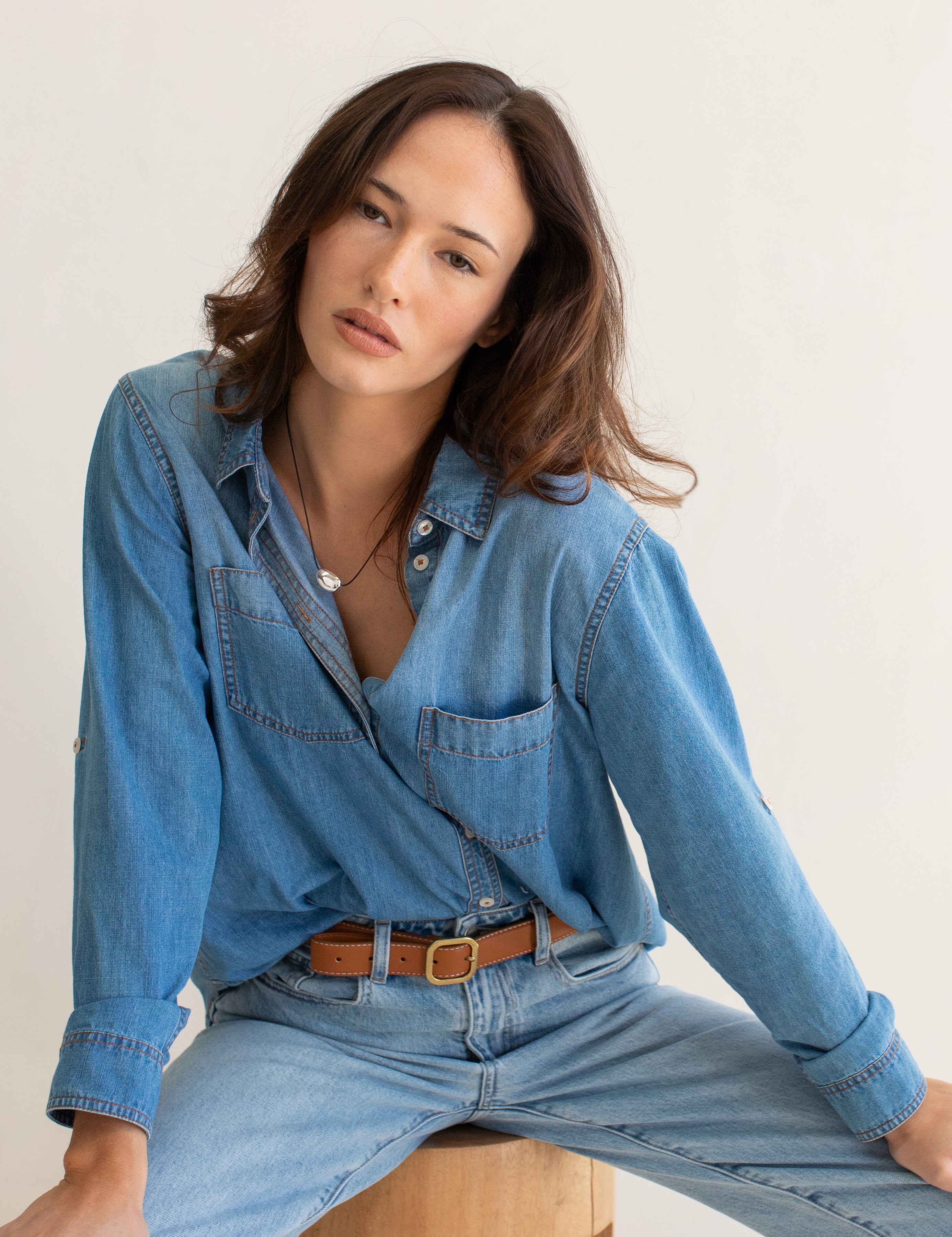 Woman wearing a blue Hut denim oversized boyfriend shirt and jeans sitting on a wooden stool against a white background
