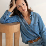Woman wearing a blue denim shirt and jeans sitting on a wooden stool against a white background