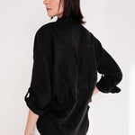 A model wearing Hut clothing black boyfriend linen shirt with an embroidered palm tree on the back, standing against a white background.