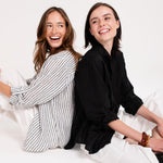 Two models back to back laughing wearing Hut Black Embroidered Palm Boyfriend Linen Shirt and Hut Black and White Stripe Boyfriend Linen Shirt
