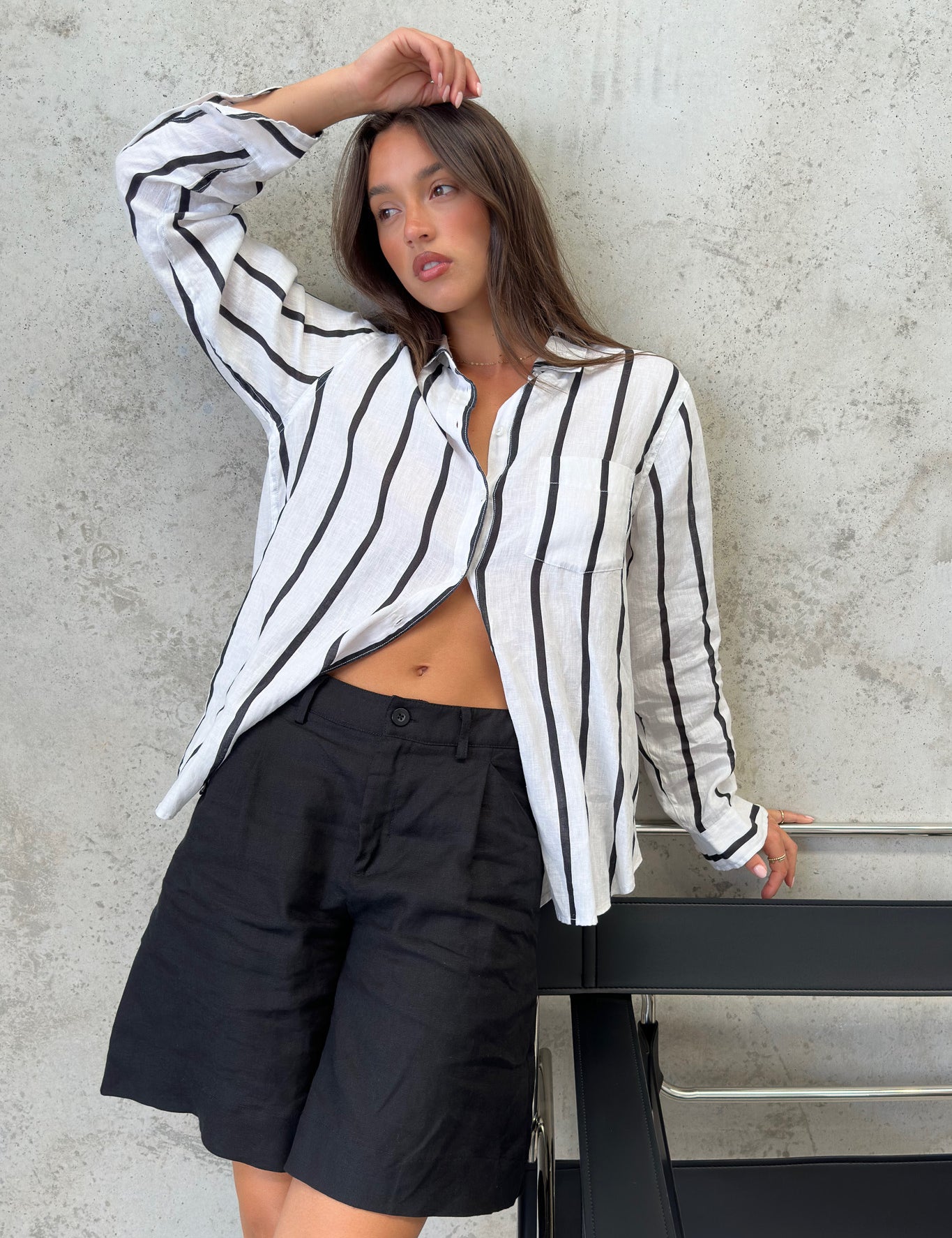 A woman wearing a black and white striped Hut boyfriend linen shirt with French seams and a scoop hem, paired with black shorts.