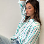 A woman sitting against a wall wearing a striped boyfriend linen shirt in green and white with rolled-up sleeves, light wash denim jeans