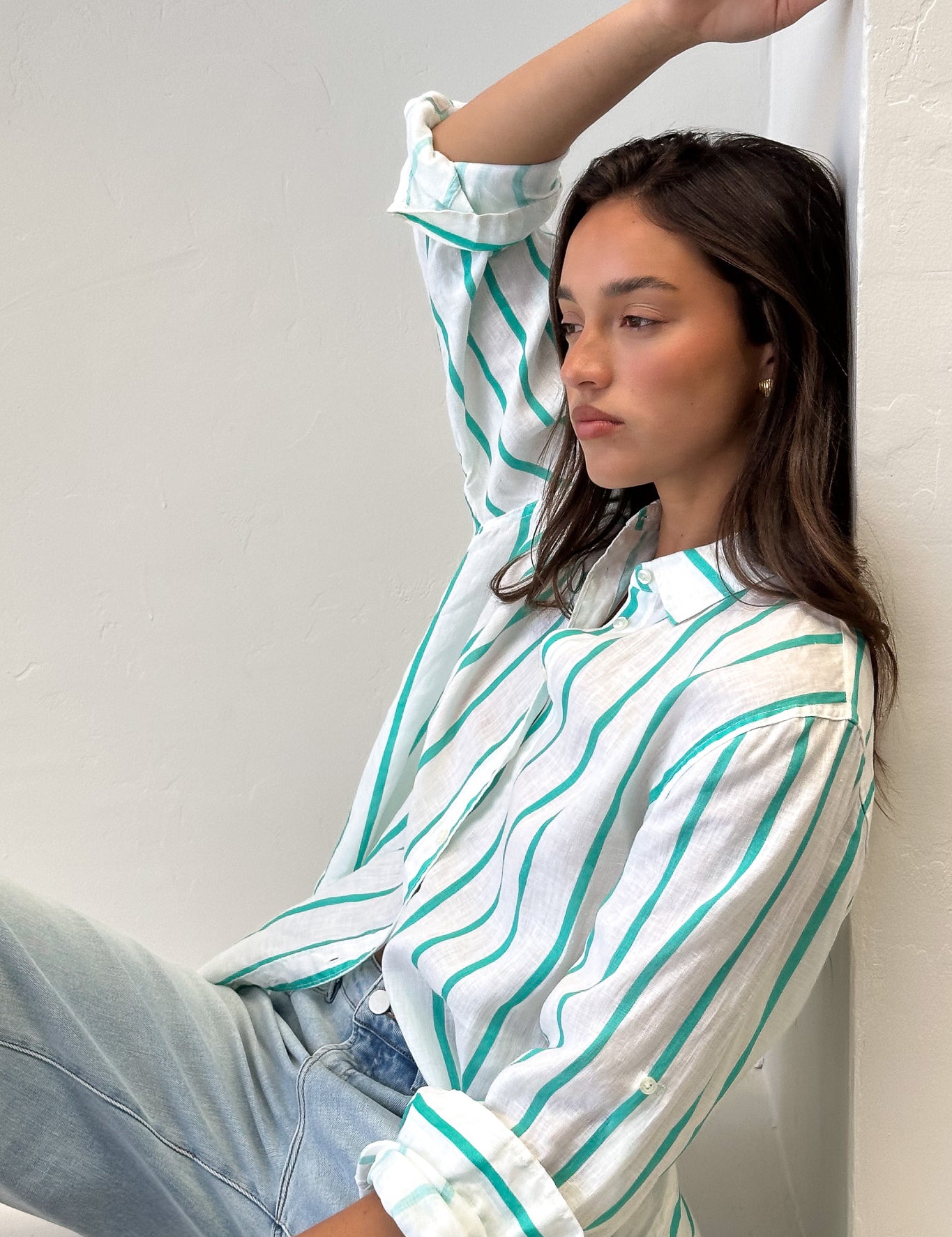 A woman sitting against a wall wearing a striped boyfriend linen shirt in green and white with rolled-up sleeves, light wash denim jeans