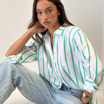Model sitting down wearing Bermuda Green Stripe Boyfriend Linen shirt with light wash denim jeans
