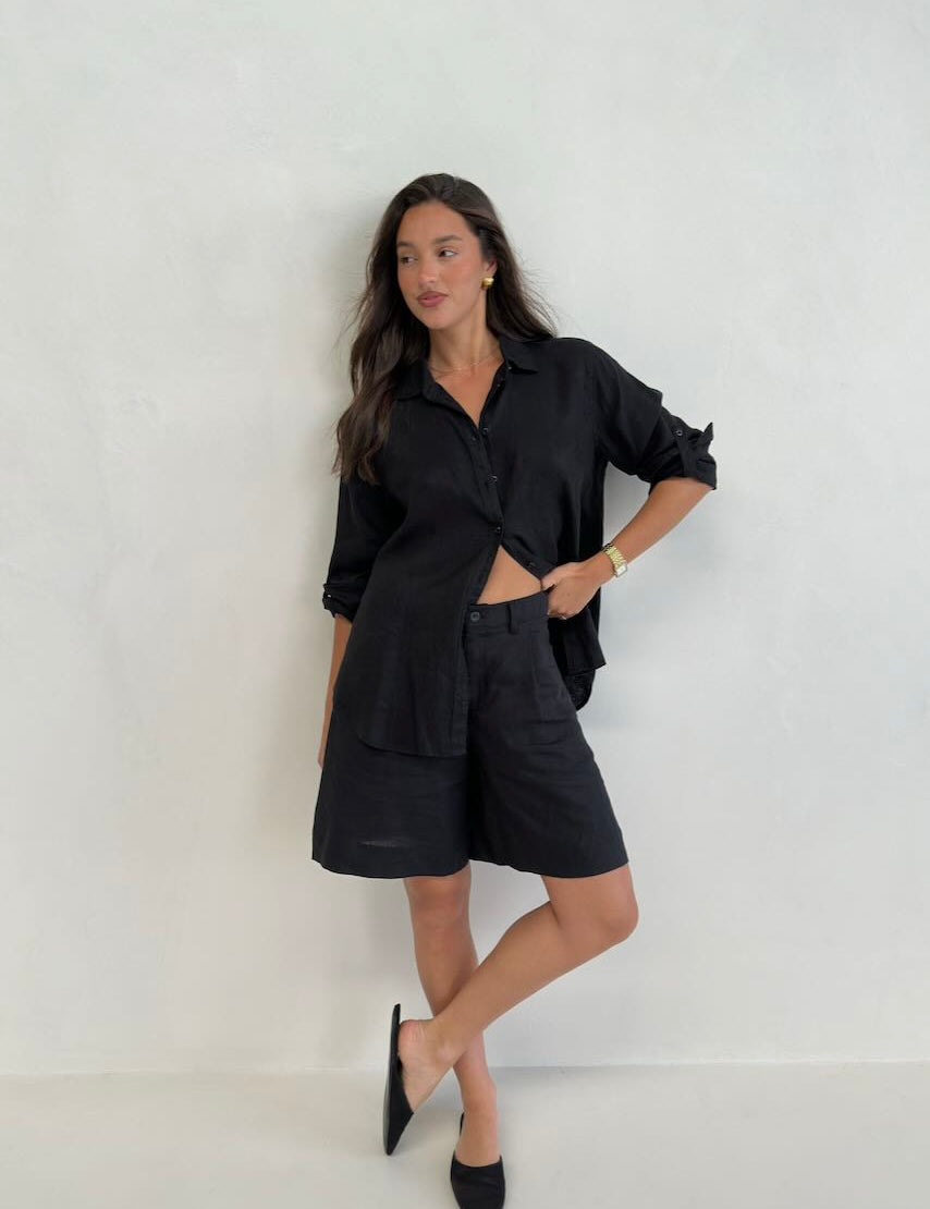 A model standing against a white background, wearing a black boyfriend linen shirt with button-up sleeves and black shorts.