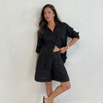 A model standing against a white background, wearing a black boyfriend linen shirt with button-up sleeves and black shorts.