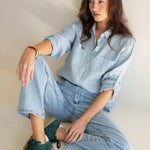 Model sitting down wearing Hut Clothing Breeze Blue Boyfriend Linen Shirt with light denim jeans and green sneakers