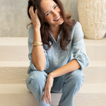 Woman sitting on a staircase wearing a light blue linen boyfriend shirt and jeans, smiling.