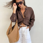 Model wearing Hut Clothing Chocolate Martini Boyfriend Linen Shirt with wind in her hair and a tan leather bag with white linen shorts