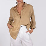A woman wearing a Hut beige linen boyfriend shirt with a collar, button-up, two chest pockets and long sleeves, paired with white trousers.