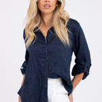 Model wearing a navy blue linen boyfriend shirt with long sleeves and white pants. The shirt has a button-up front and a scooped hem.