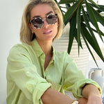 A woman wearing a green linen boyfriend shirt with rolled-up sleeves, paired with white shorts, sitting beside a potted plant.