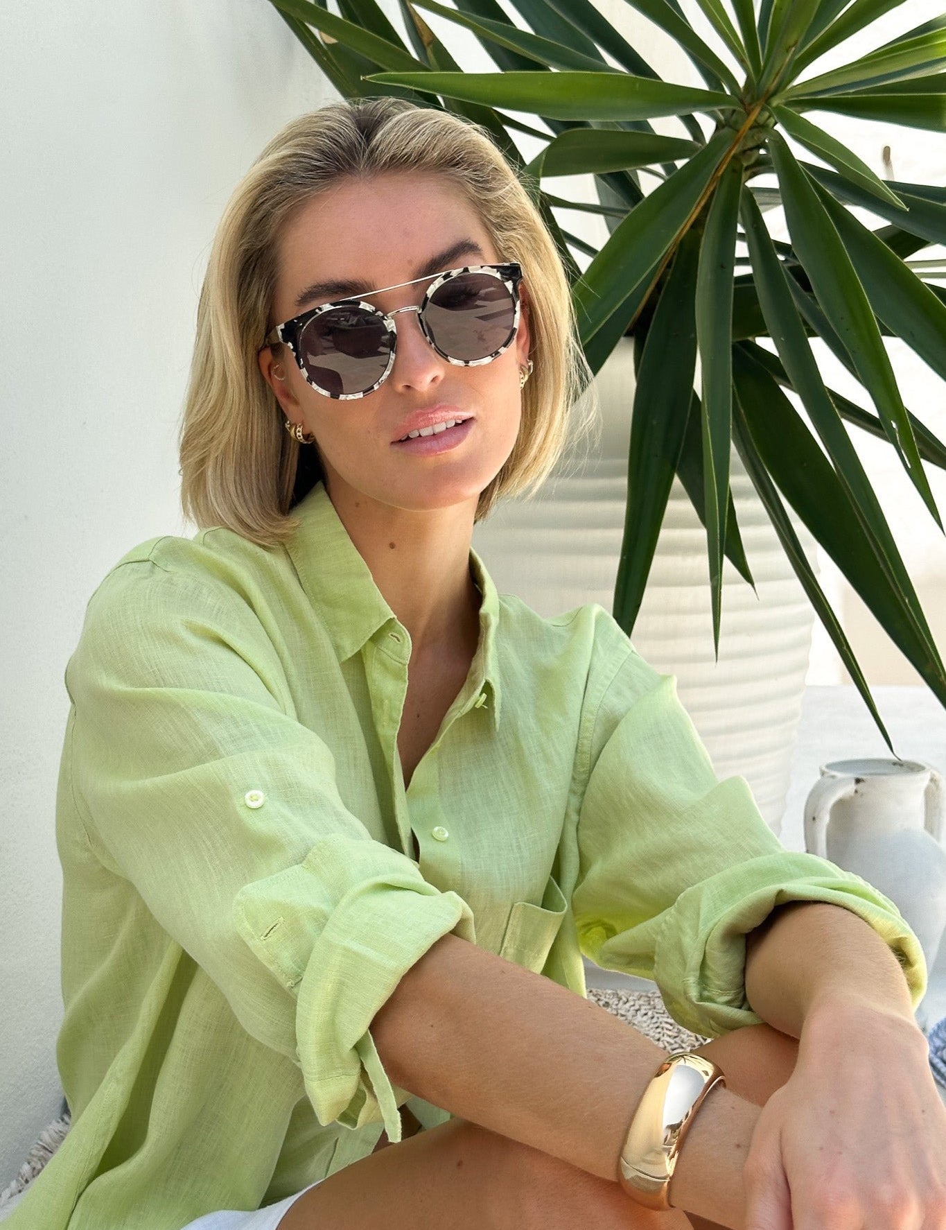 A woman wearing a green linen boyfriend shirt with rolled-up sleeves, paired with white shorts, sitting beside a potted plant.