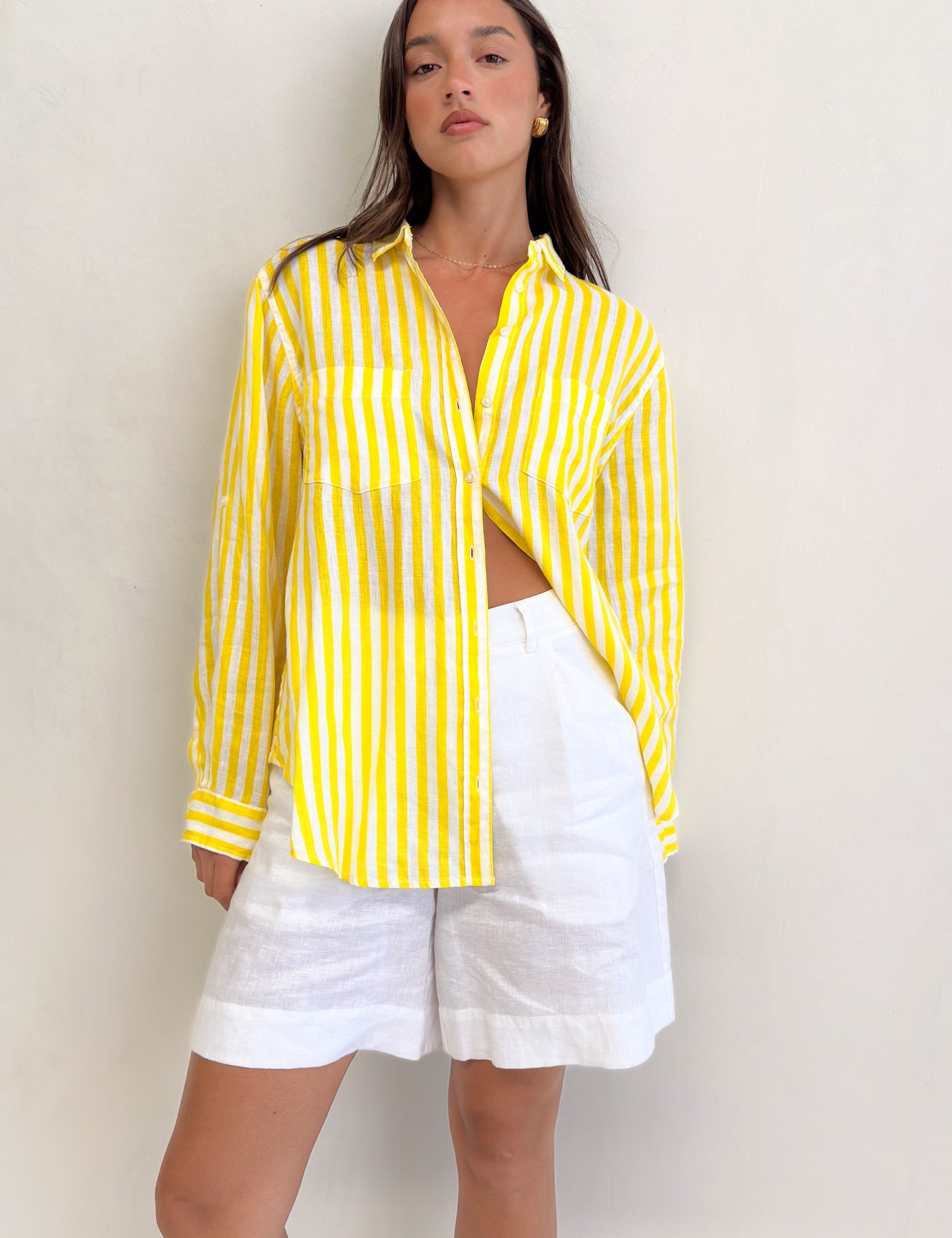A woman wearing a Hut Clothing yellow and white striped linen boyfriend shirt with long sleeves and two chest pockets, paired with white shorts.