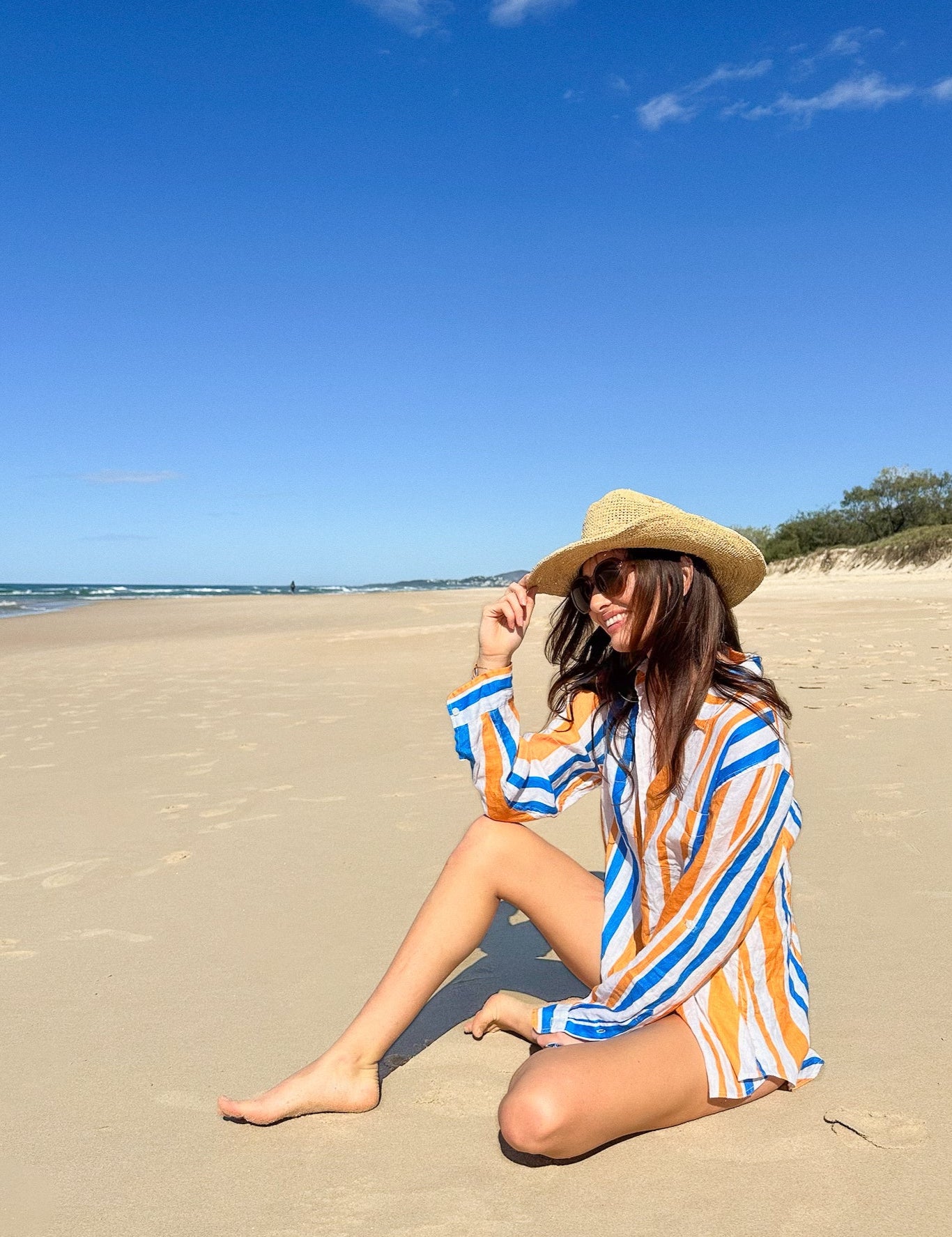 Woman sitting on a sandy beach wearing an orange and blue striped linen oversized shirt and hat.