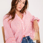 A woman sitting and wearing a Hut red and white striped boyfriend linen shirt with button-up sleeves and a scoop hem, paired with blue jeans.
